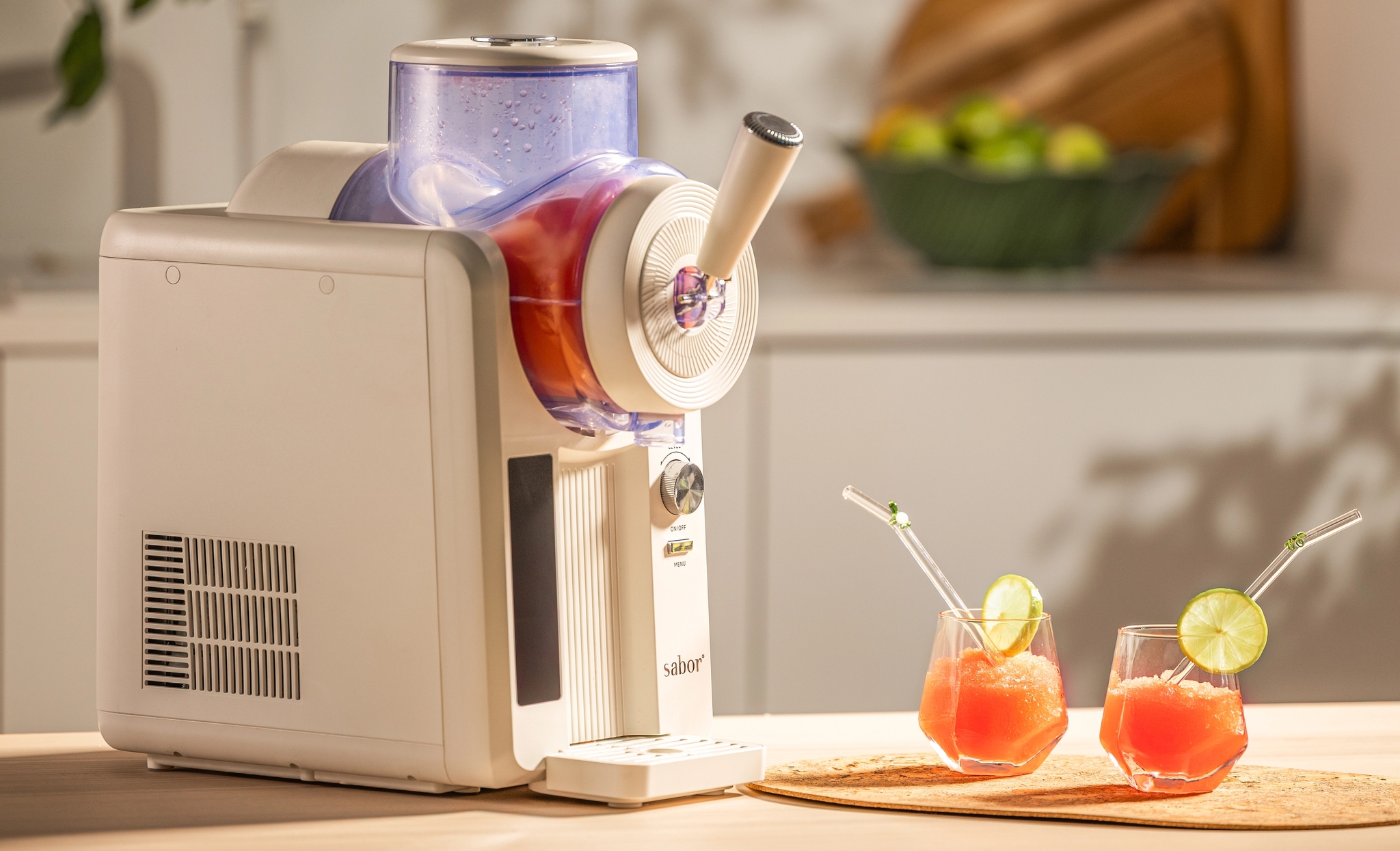 Valkoinen slushie-kone keittiön tiskillä kahden punaisen slushiedrinkin vieressä, koristeltuna limenviipaleilla ja pilleillä.