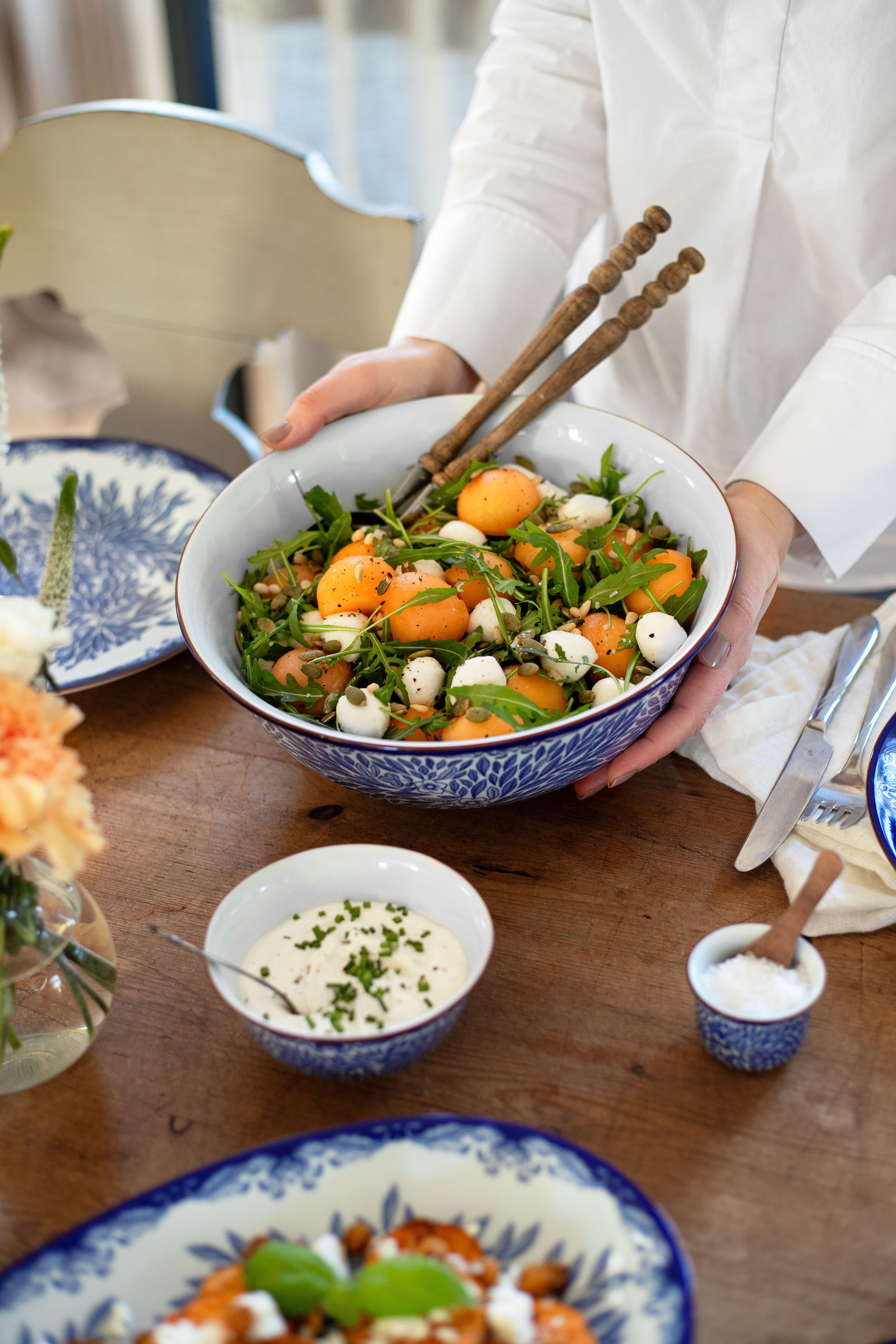 Person serverar en somrig sallad med aprikoser, ruccola och mozzarella i en blåvit skål på ett dukat träbord.