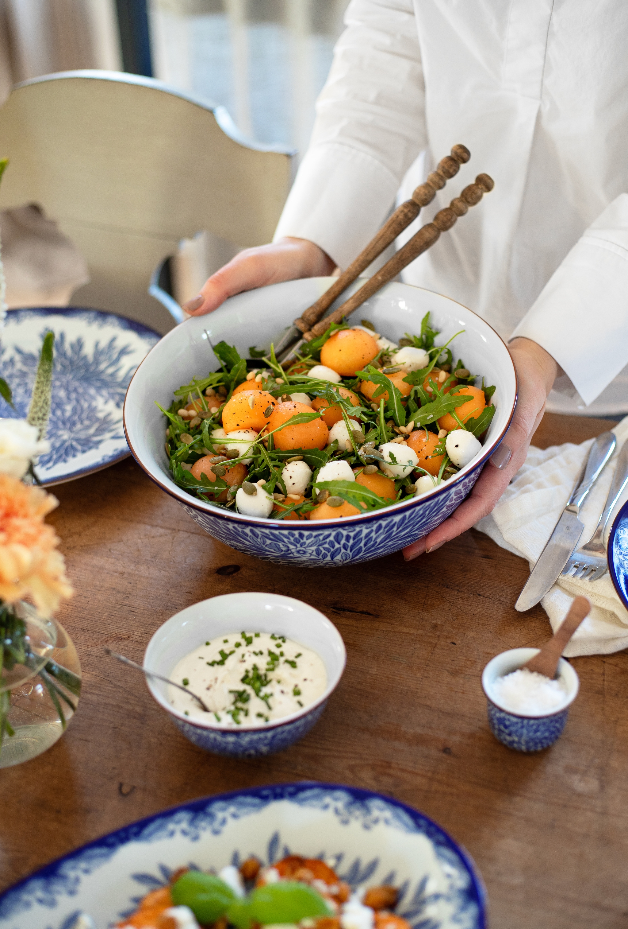 Person som serverer en stor sommersalat med aprikoser, ruccola og mozzarella i en blåmønstret bolle på et rustikt trebord.
