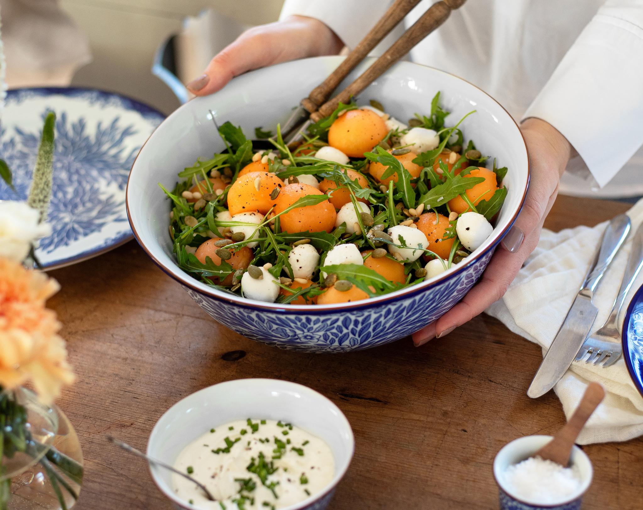 Person serverar en somrig sallad med aprikoser, ruccola och mozzarella i en blåvit skål på ett dukat träbord.