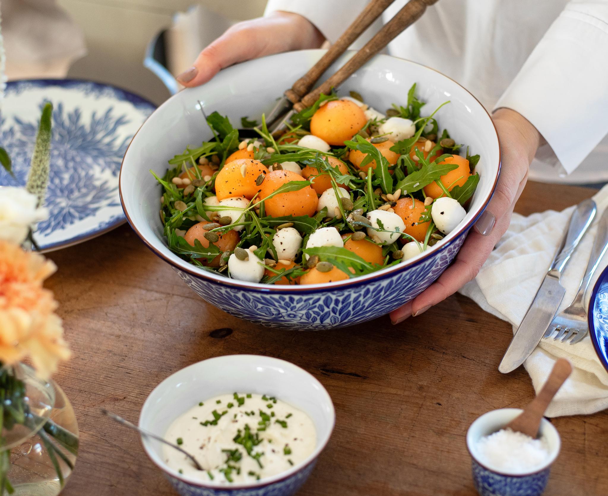 Person som serverer en stor sommersalat med aprikos, ruccola og mozzarella i en blåmønstret bolle på et dekket trebord.
