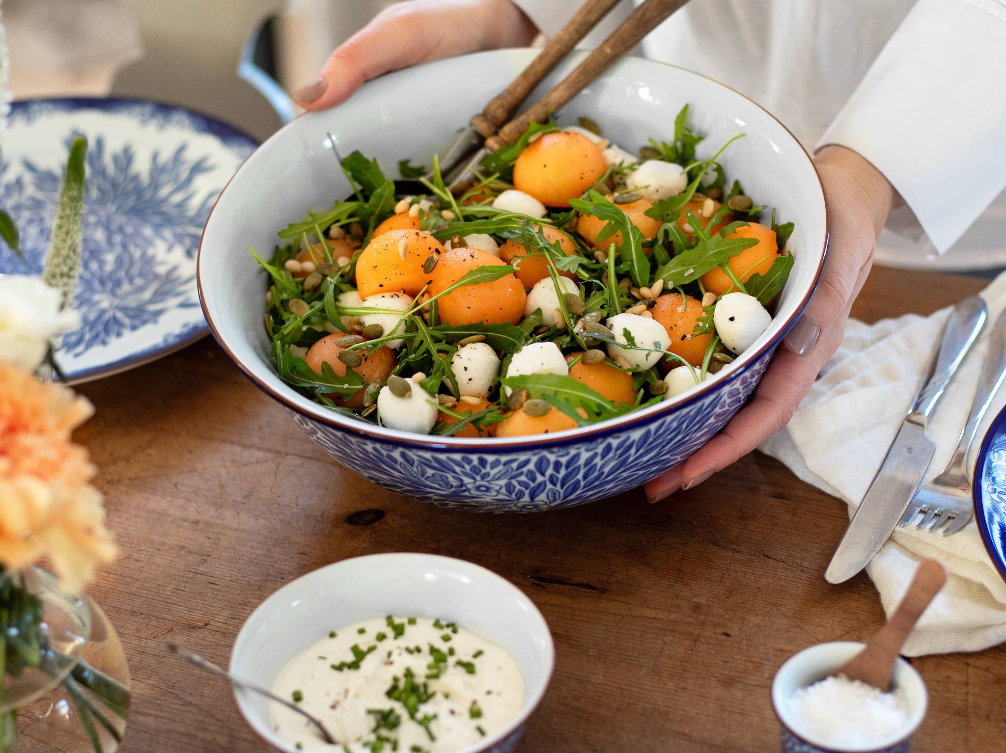 Person serverar en stor skål fräsch sallad med aprikoser, ruccola och mozzarella på ett dukat träbord.