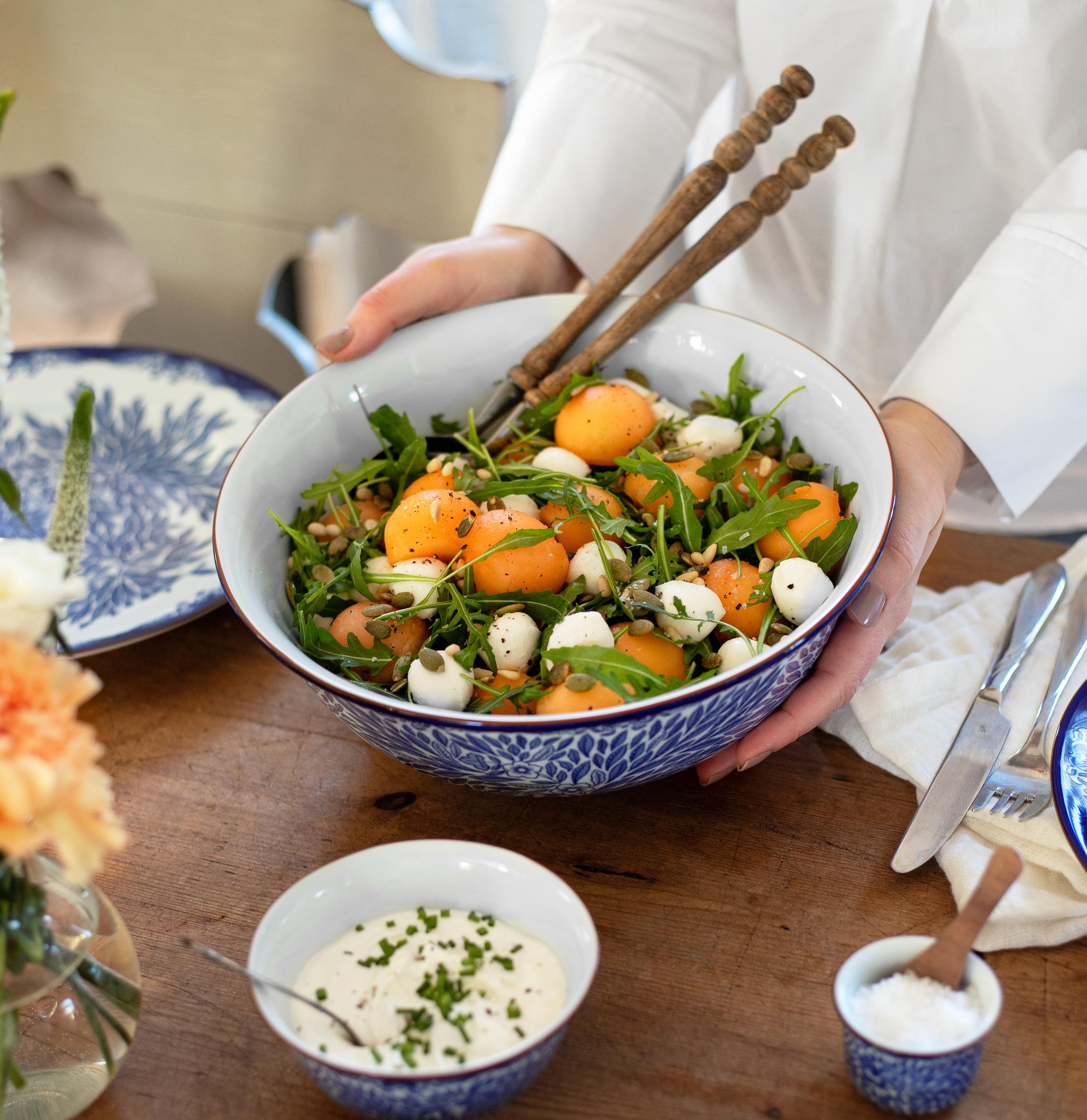 Person serverer en stor sommersalat med aprikoser, ruccola og mozzarella i en blåmønstret bolle på et dekket trebord.