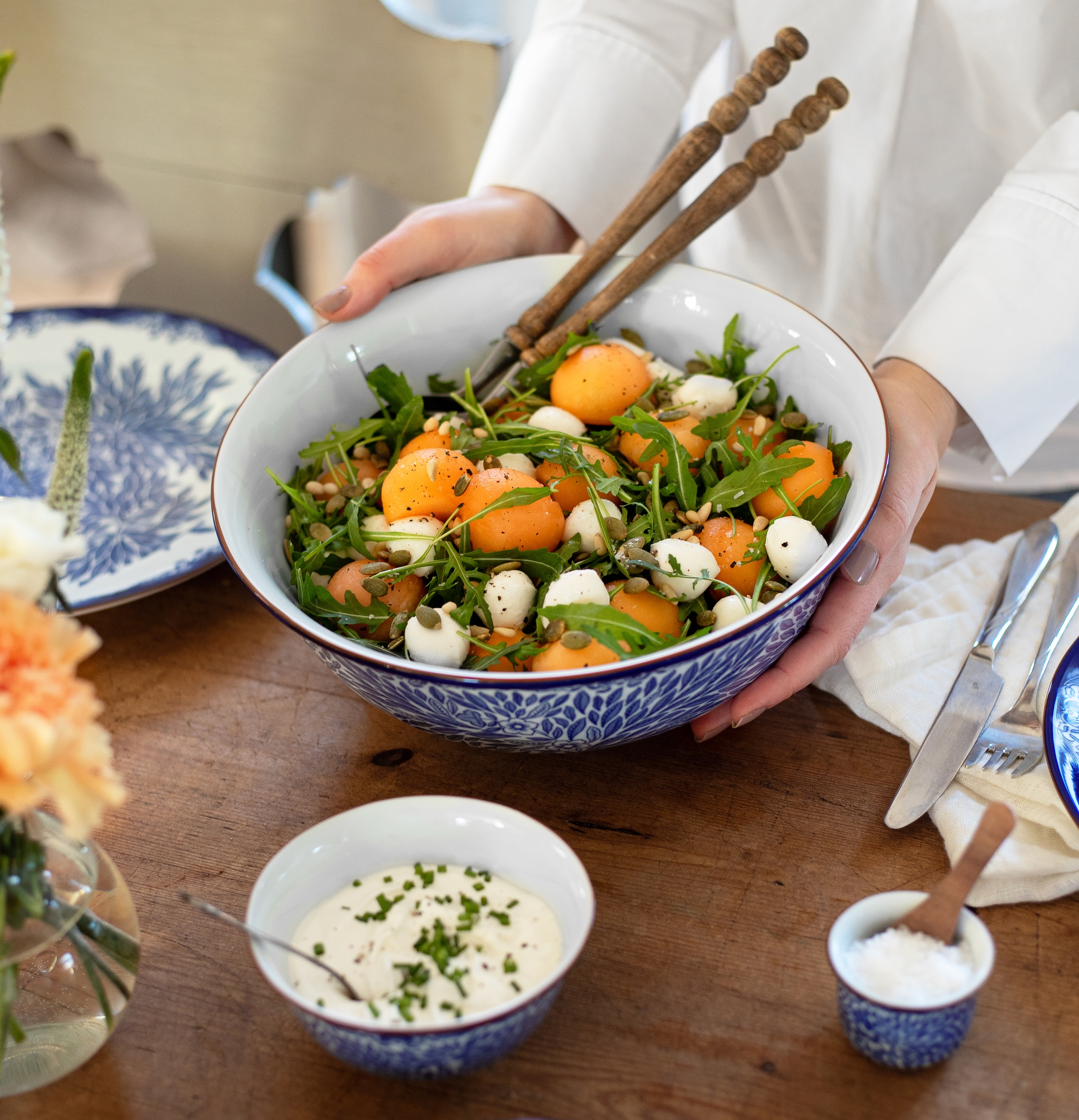 Person som serverer en stor sommersalat med aprikoser, ruccola og mozzarella på et rustikt trebord dekket til måltid.