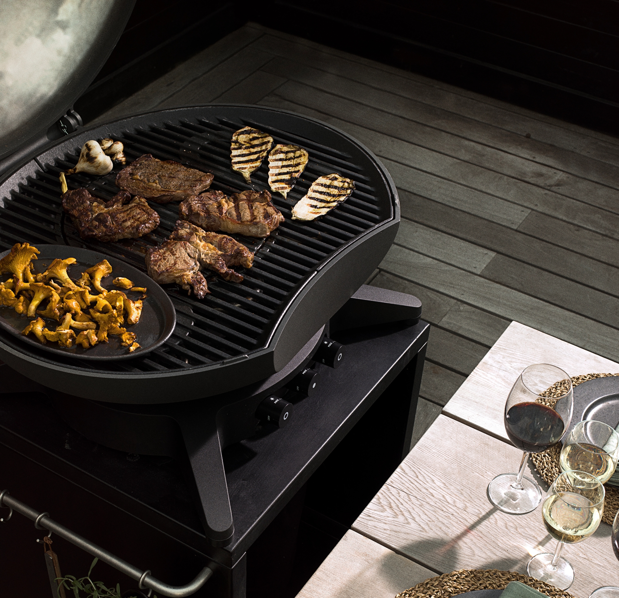En grill med grillat kött, ost, svamp och vitlök, bredvid ett dukat matbord med vin på en träaltan