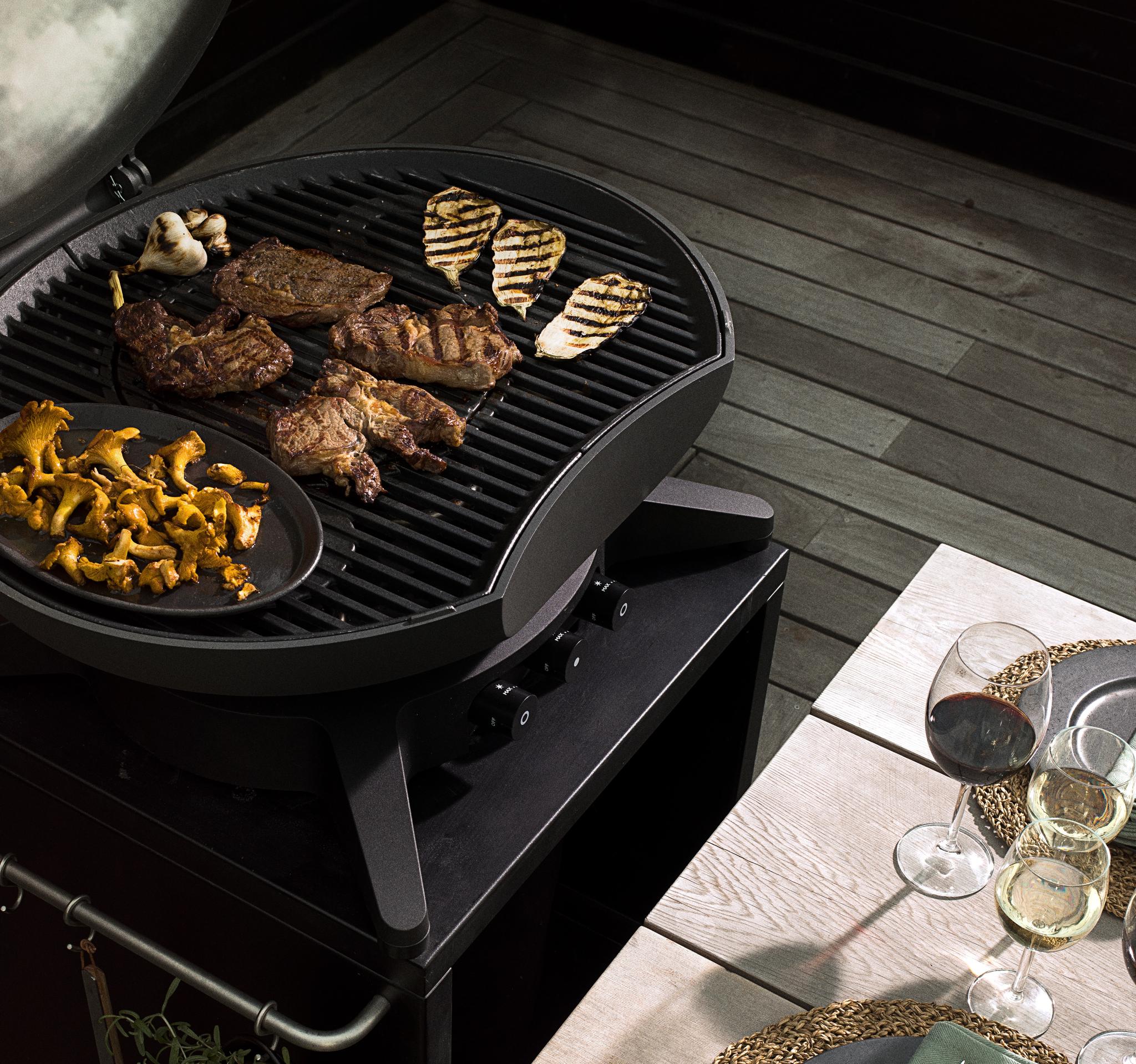 En grill med grillat kött, ost, svamp och vitlök, bredvid ett dukat matbord med vin på en träaltan