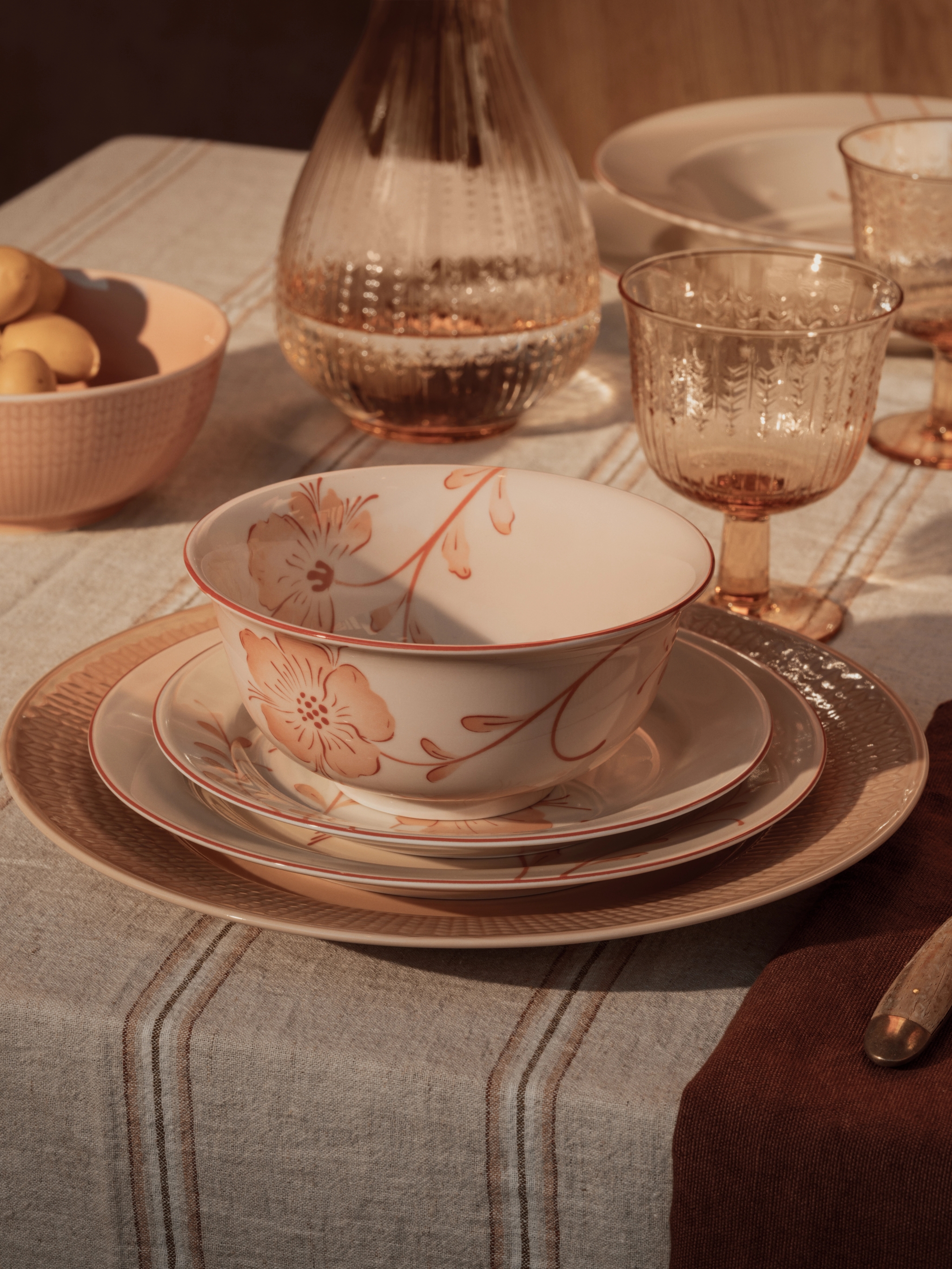 En borddekking i varme toner med en blomstrete bolle og stablede tallerkener, ravfargede glass og en bolle med sitroner på en stripete duk.