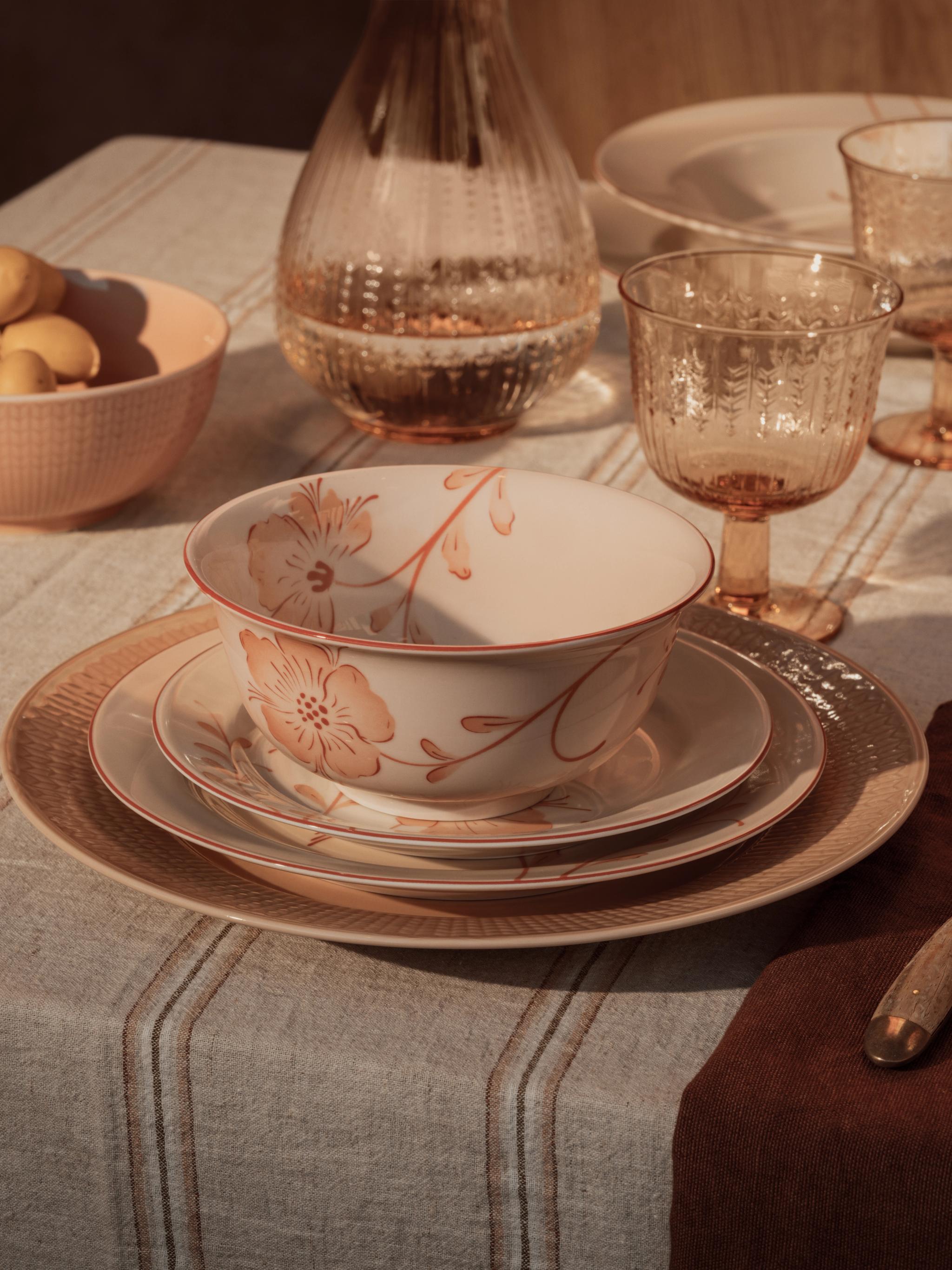 En borddekking i varme toner med en blomstrete bolle og stablede tallerkener, ravfargede glass og en bolle med sitroner på en stripete duk.