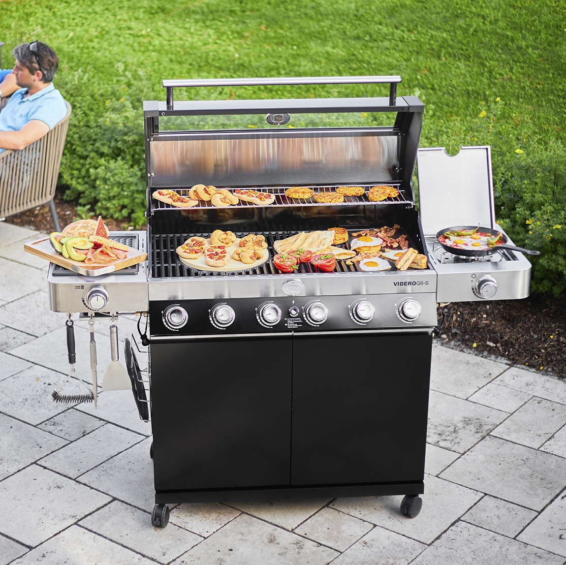 A black and stainless steel gas grill cooking a diverse spread of food, including pizzas, eggs, and bacon, on an outdoor patio.