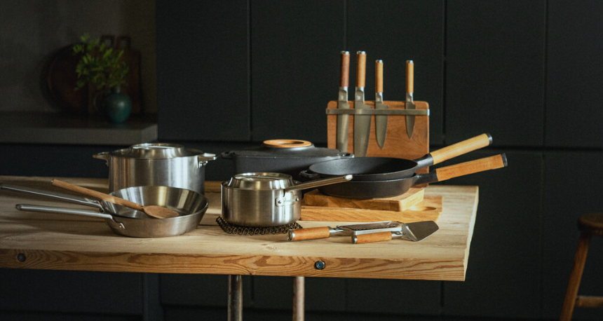 Moderne kjøkkenbenk med gryter, stekepanner og knivsett i rustfritt stål og trehåndtak.