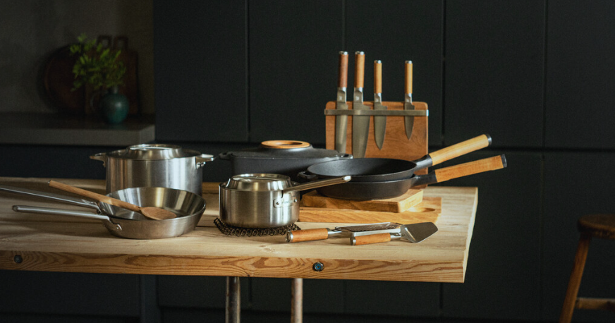 Moderne kjøkkenbenk med gryter, stekepanner og knivsett i rustfritt stål og trehåndtak.