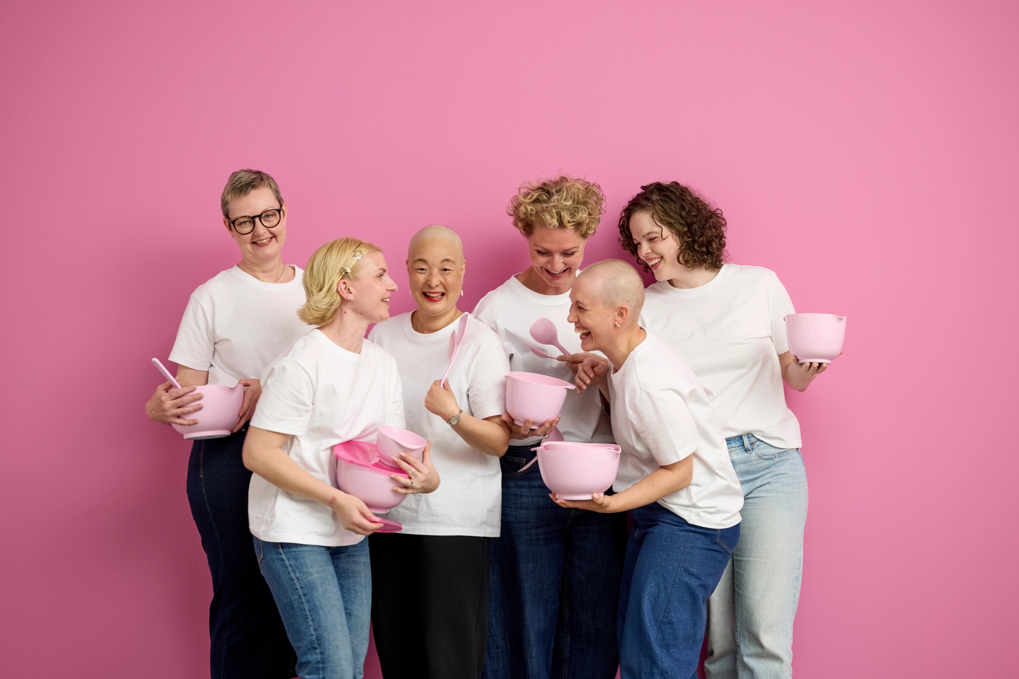 Sex leende kvinnor i vita tröjor håller rosa skålar och köksredskap mot en rosa bakgrund.