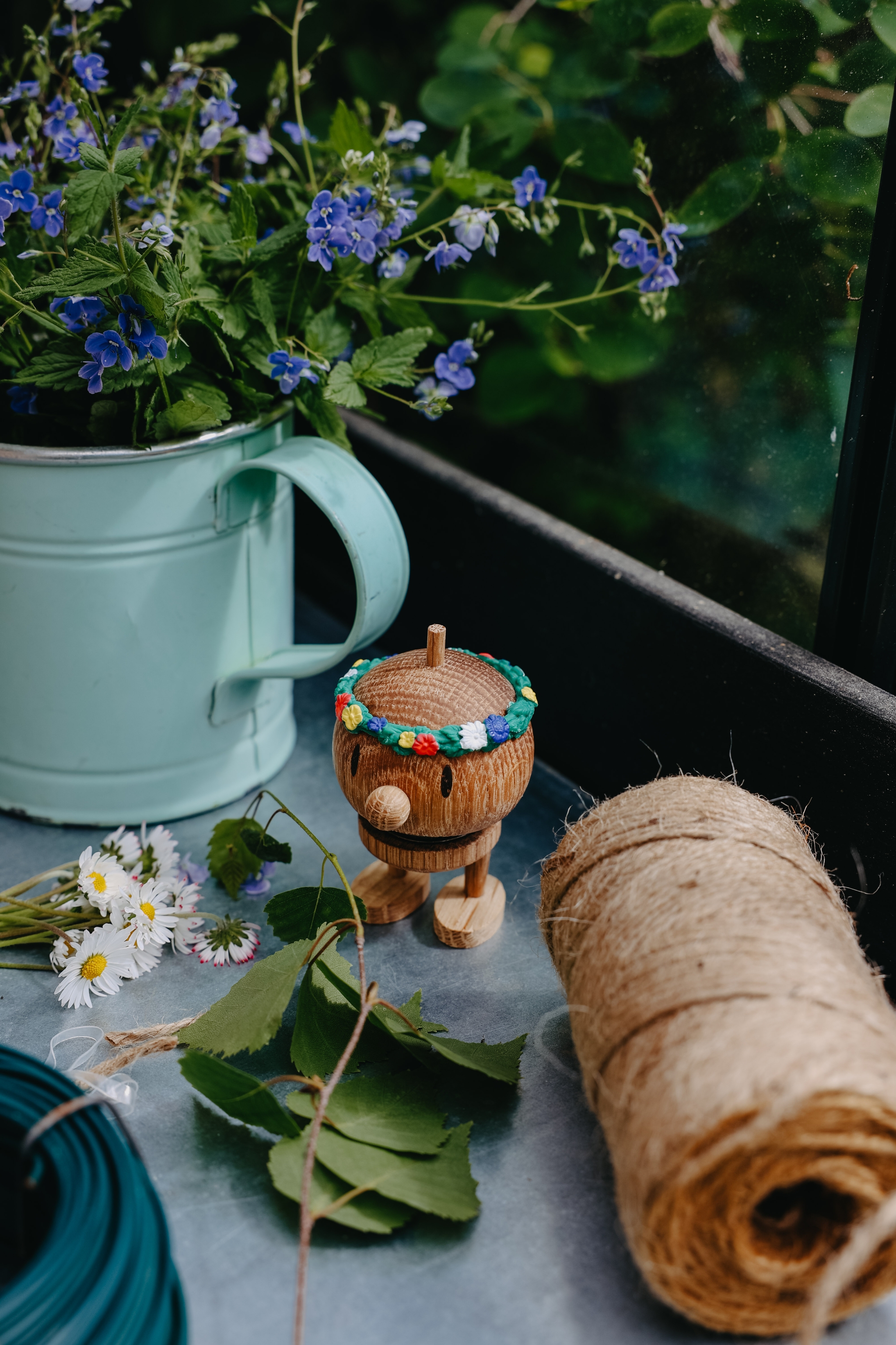 Somrig stilleben med blå blommor i en ljusgrön kanna, en liten träfigur med midsommarkrans och trädgårdssnöre på ett bord.