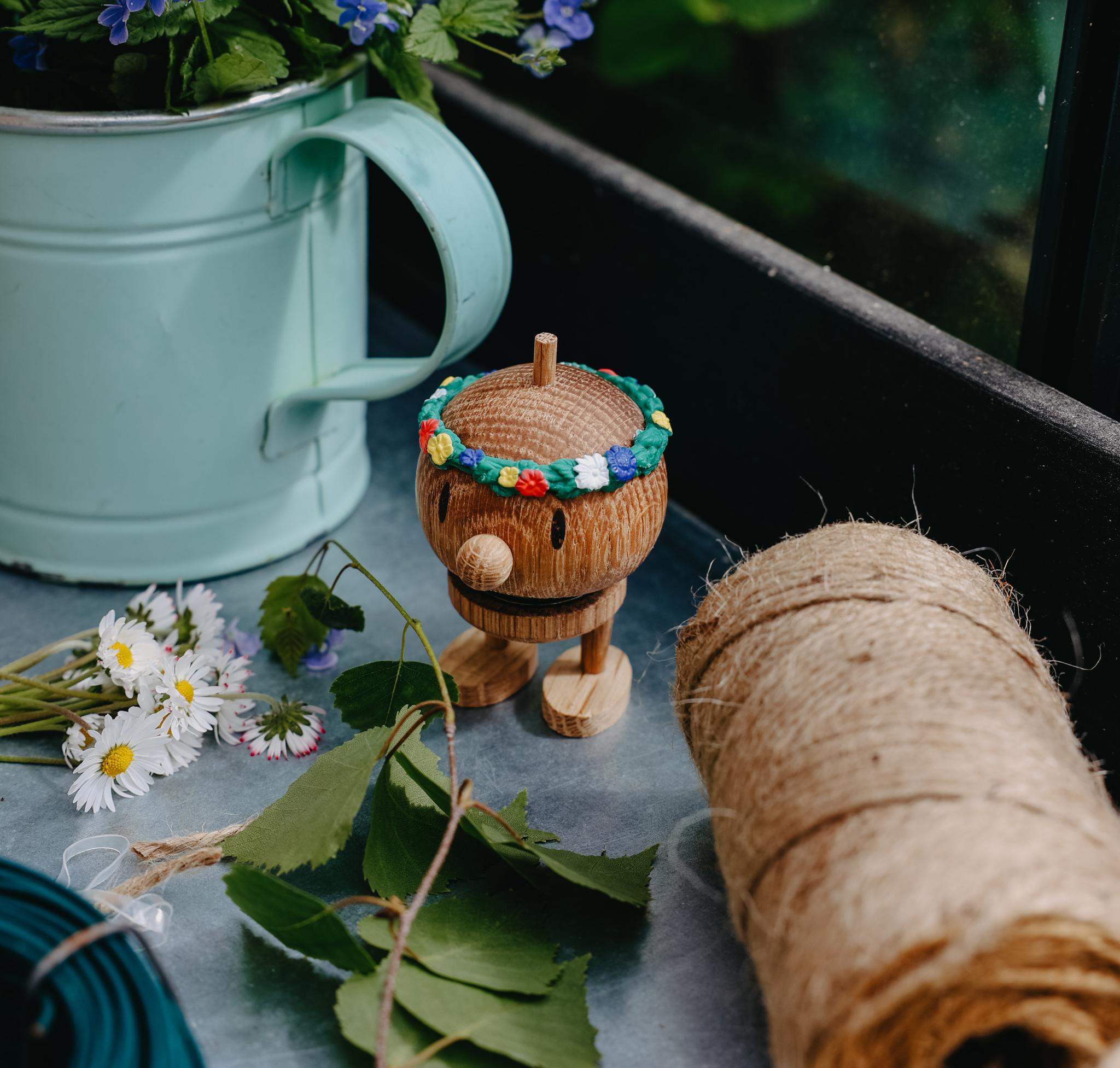 Somrig trädgårdsscen med blå blommor i en ljusgrön kanna, en liten träfigur med blomkrans och trädgårdssnöre på ett bord.