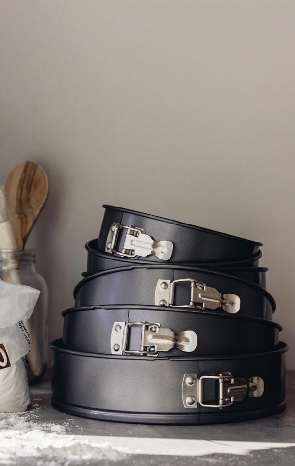 Pino mustia irtopohjaisia kakkuvuokia, ympärillä jauhoja ja puinen lusikka.