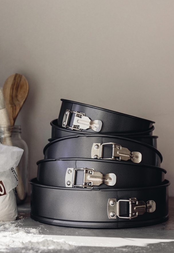 Pino mustia irtopohjaisia kakkuvuokia, ympärillä jauhoja ja puinen lusikka.