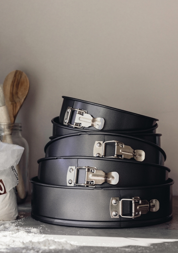 Pino mustia irtopohjaisia kakkuvuokia, ympärillä jauhoja ja puinen lusikka.