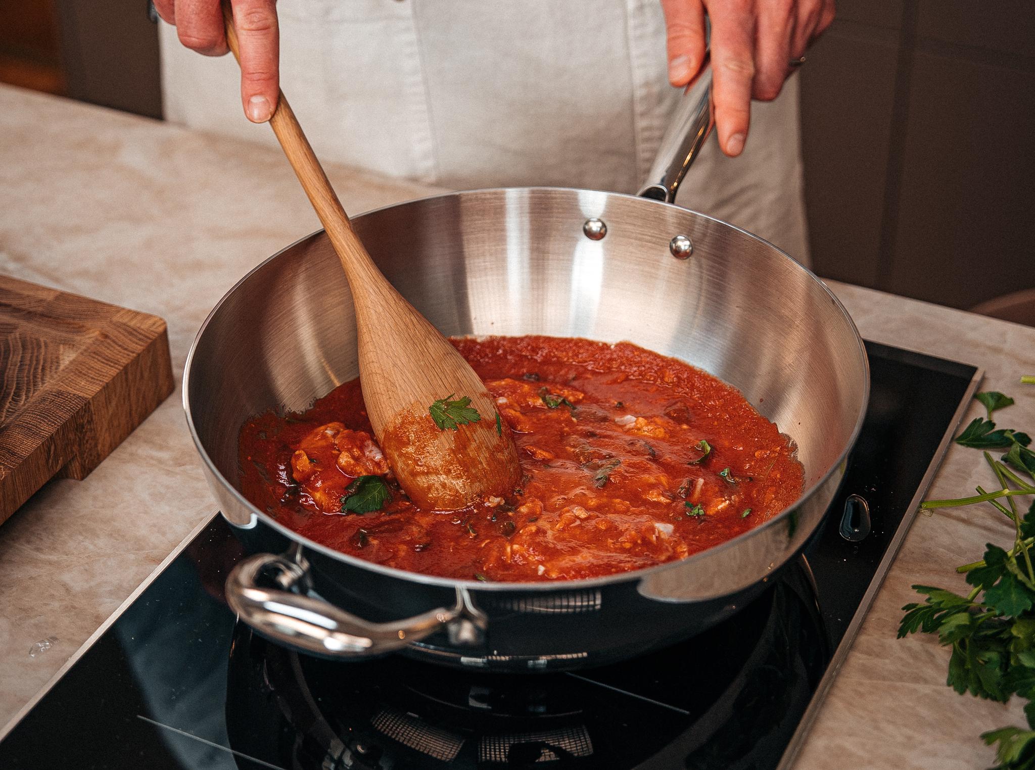 Person som rör om en tomatbaserad pastasås i en stor stekpanna på spisen.