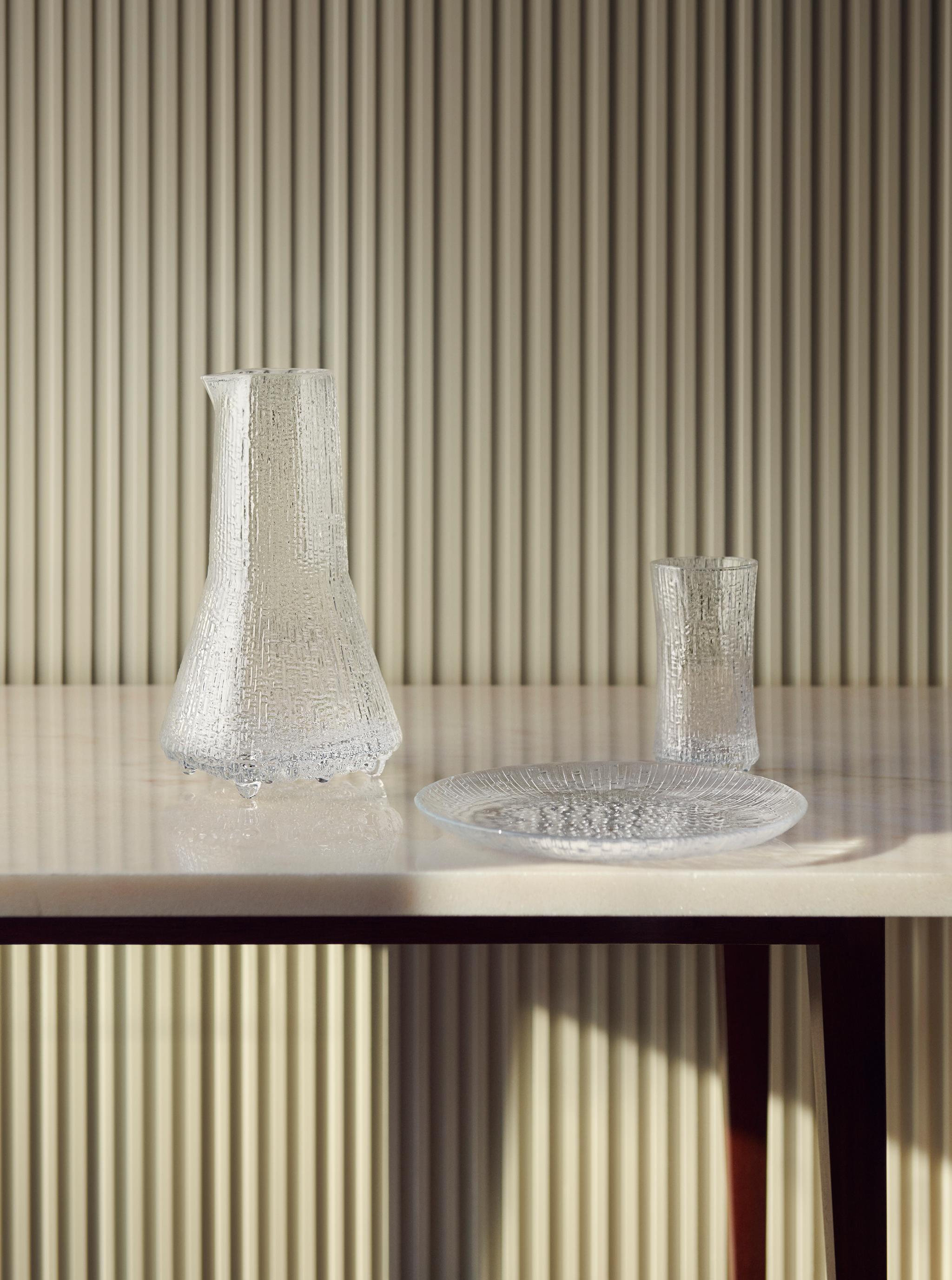 Klar glasskaraffel, drikkeglass og tallerken på et lyst bord foran en rillet vegg.