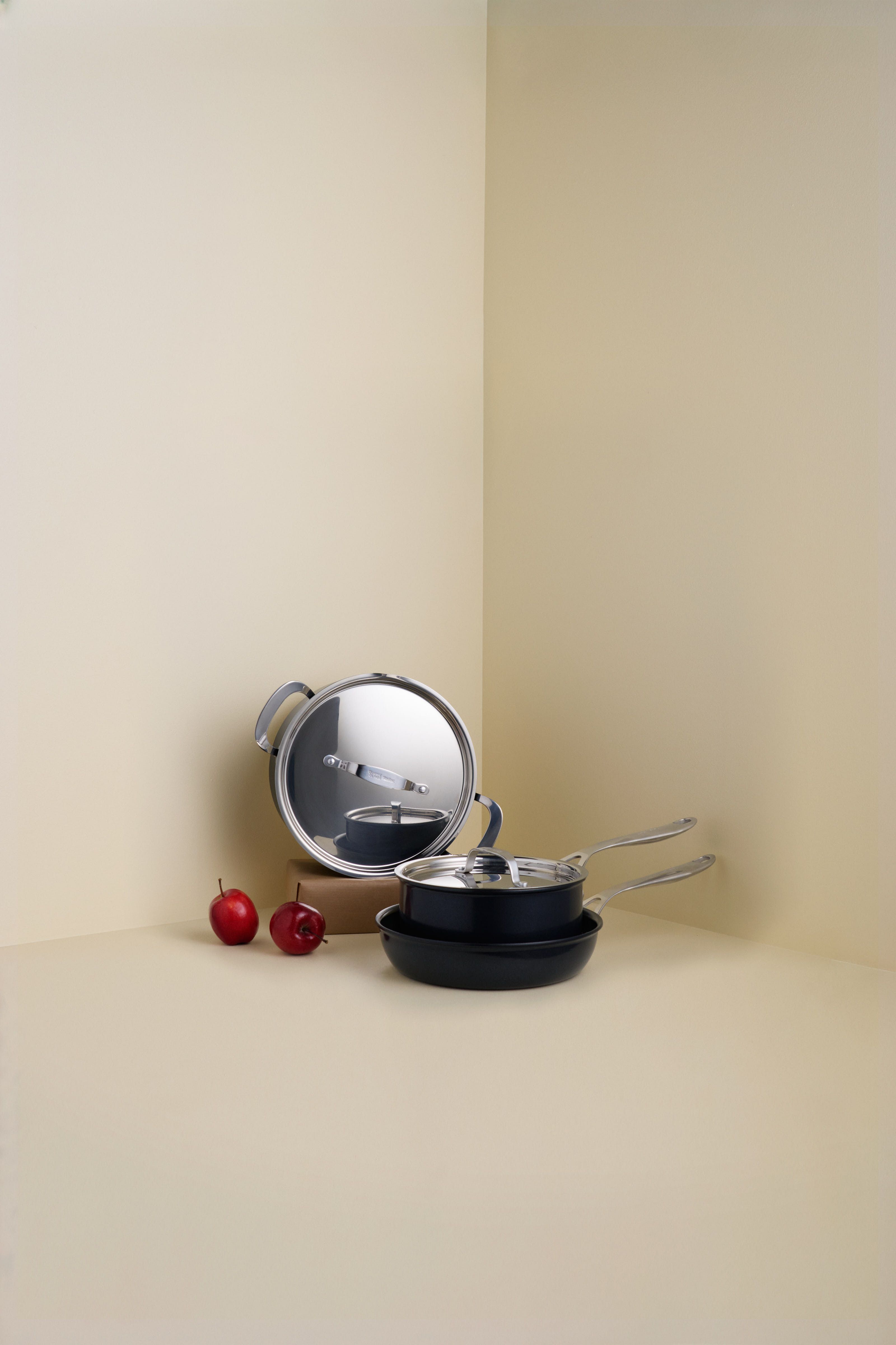 Et sett med svarte gryter og panner med reflekterende lokk, og to røde epler, i et beige hjørne.