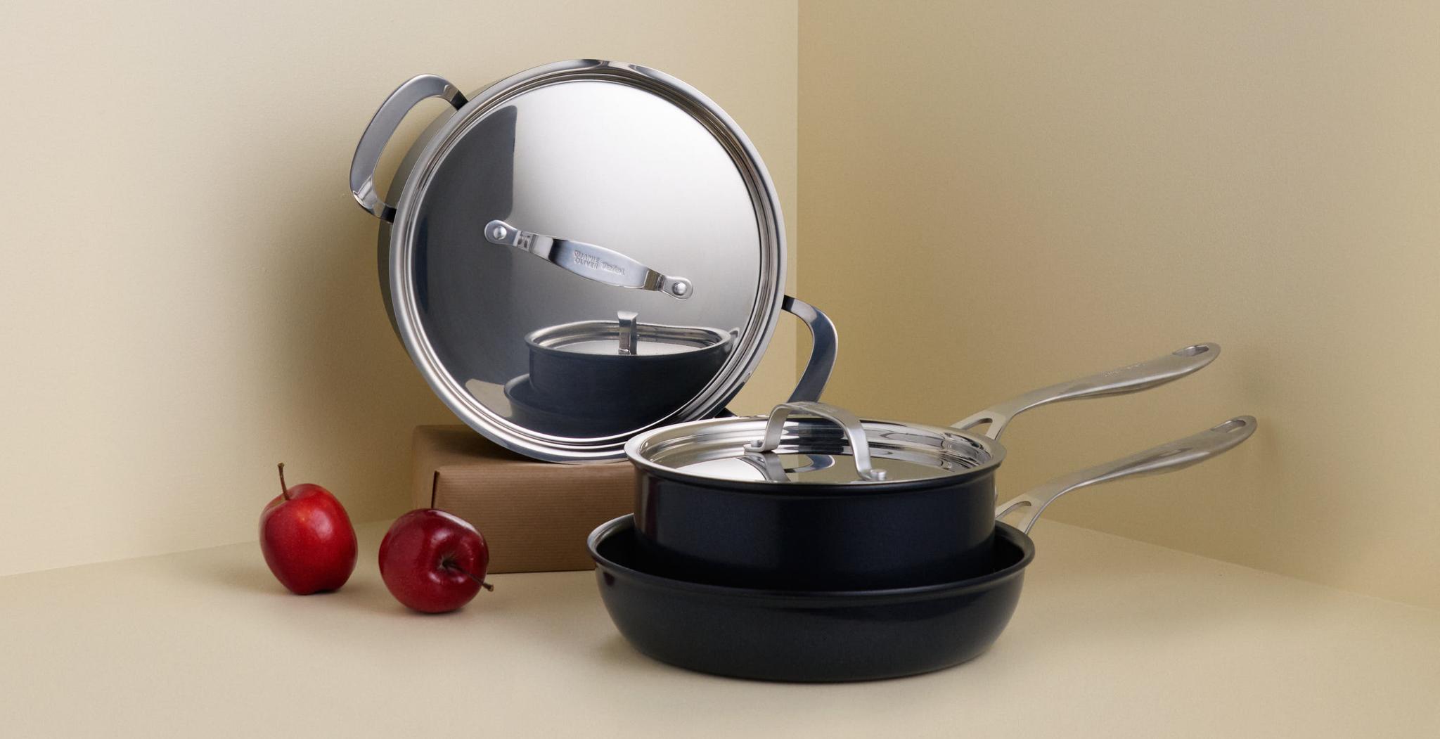 En uppsättning svarta köksredskap med glänsande silverlock och handtag, och två röda äpplen, arrangerade i ett ljust hörn.