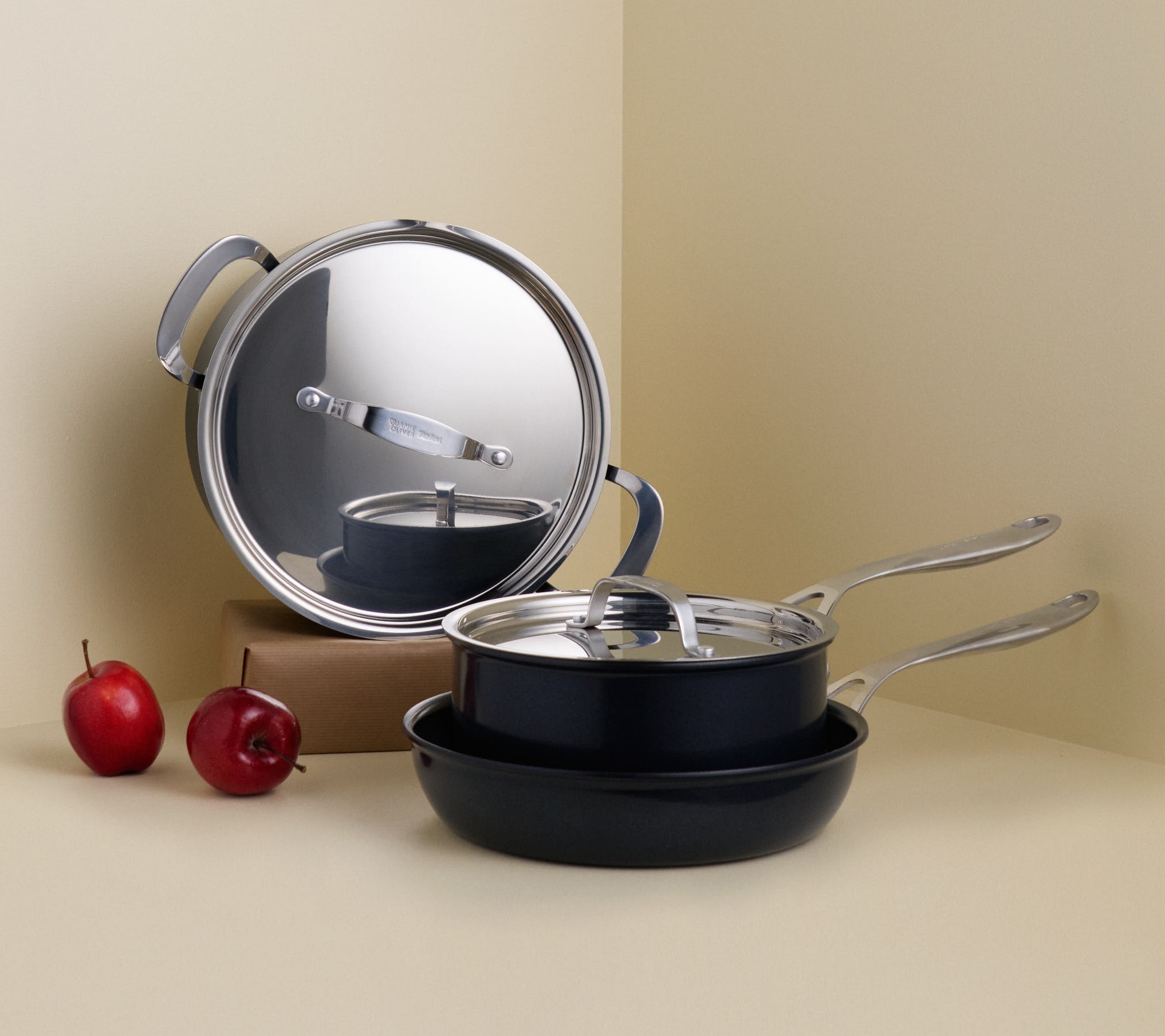 Mustat kattilat ja pannut hopeanvärisillä yksityiskohdilla ja kaksi punaista omenaa ovat vaaleanbeigessä huoneennurkassa.