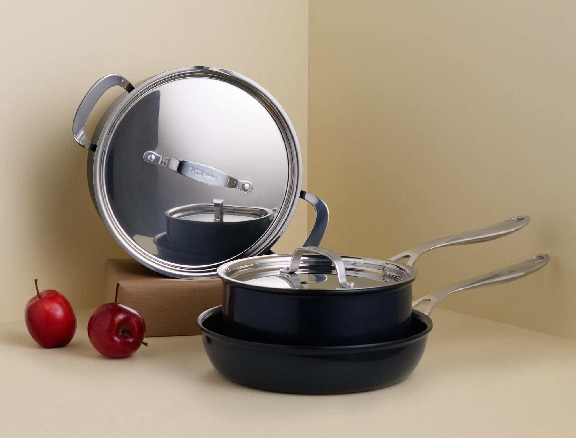 Mustat kattilat ja pannut hopeanvärisillä yksityiskohdilla ja kaksi punaista omenaa ovat vaaleanbeigessä huoneennurkassa.