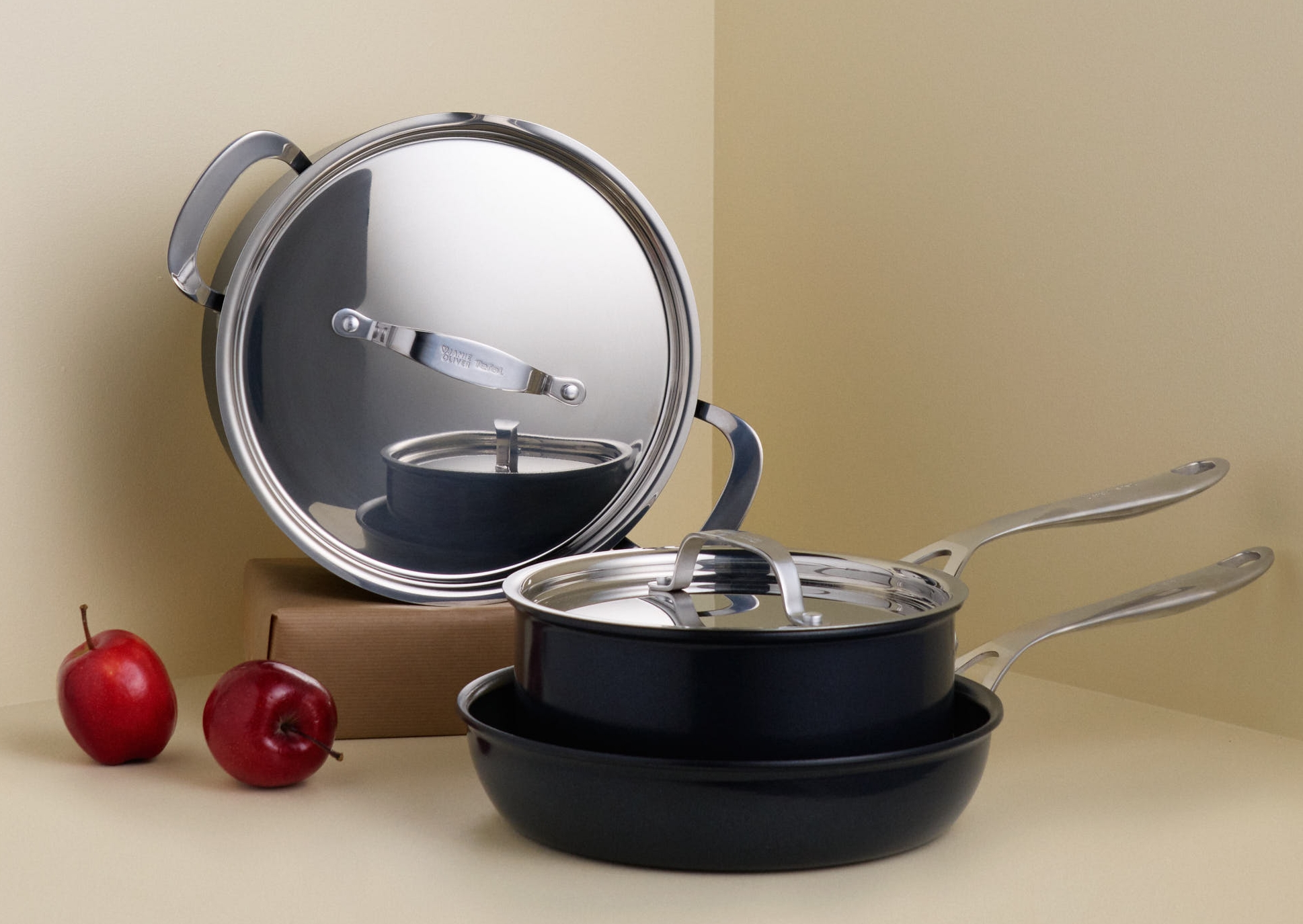 En uppsättning svarta köksredskap med glänsande silverlock och handtag, och två röda äpplen, arrangerade i ett ljust hörn.
