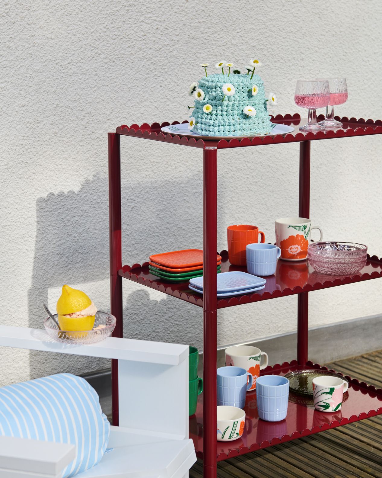 Punaisella simpukankuorisella hyllyllä on värikkäitä astioita ja sininen virkattu päivänkakkarakakku, ja lähellä valkoisella pöydällä on sitruunajälkiruoka.