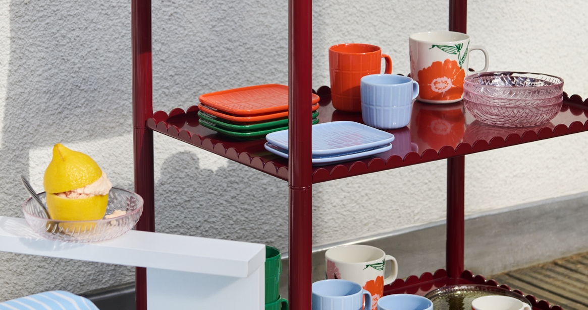 Punaisella simpukankuorisella hyllyllä on värikkäitä astioita ja sininen virkattu päivänkakkarakakku, ja lähellä valkoisella pöydällä on sitruunajälkiruoka.