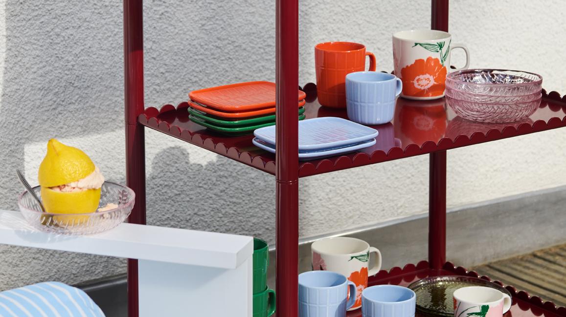 Punaisella simpukankuorisella hyllyllä on värikkäitä astioita ja sininen virkattu päivänkakkarakakku, ja lähellä valkoisella pöydällä on sitruunajälkiruoka.
