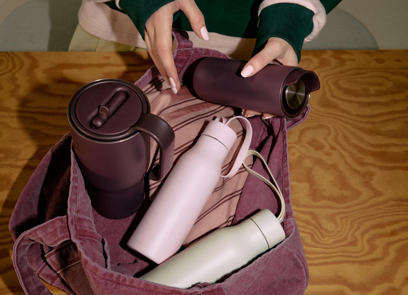 Person packar termosmuggar och återanvändbara flaskor i en rosa tygväska på ett träbord.