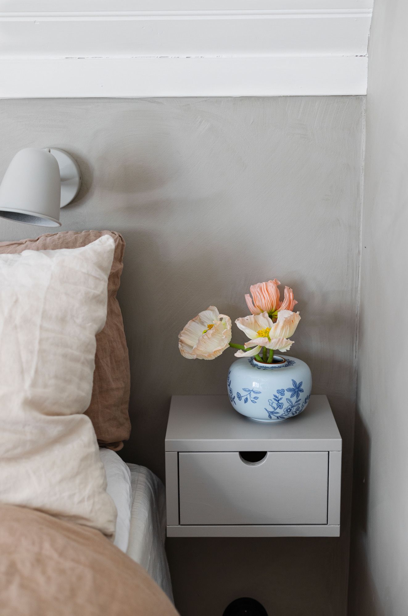 A bed with a linen pillow and duvet, a gray floating nightstand holding a blue and white vase with pale poppies, and a gray wall lamp.
