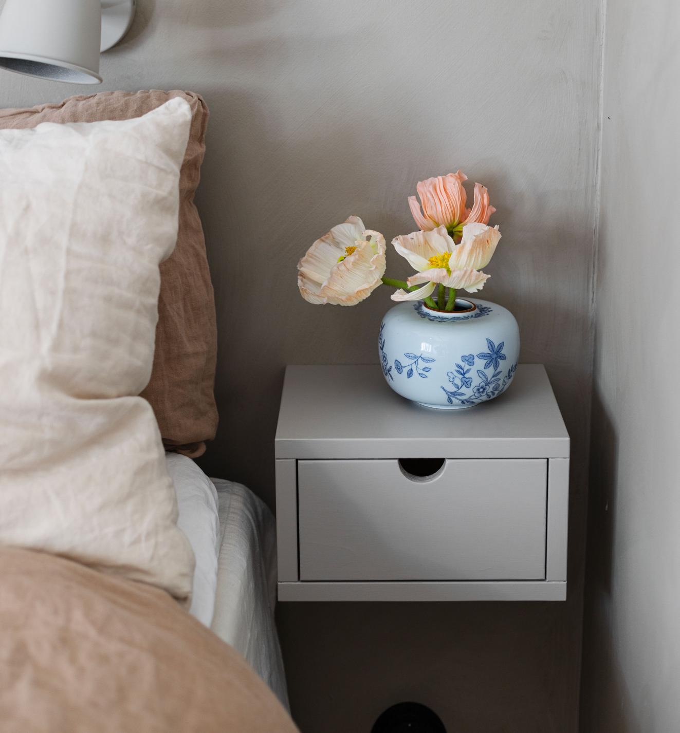 A bed with a linen pillow and duvet, a gray floating nightstand holding a blue and white vase with pale poppies, and a gray wall lamp.