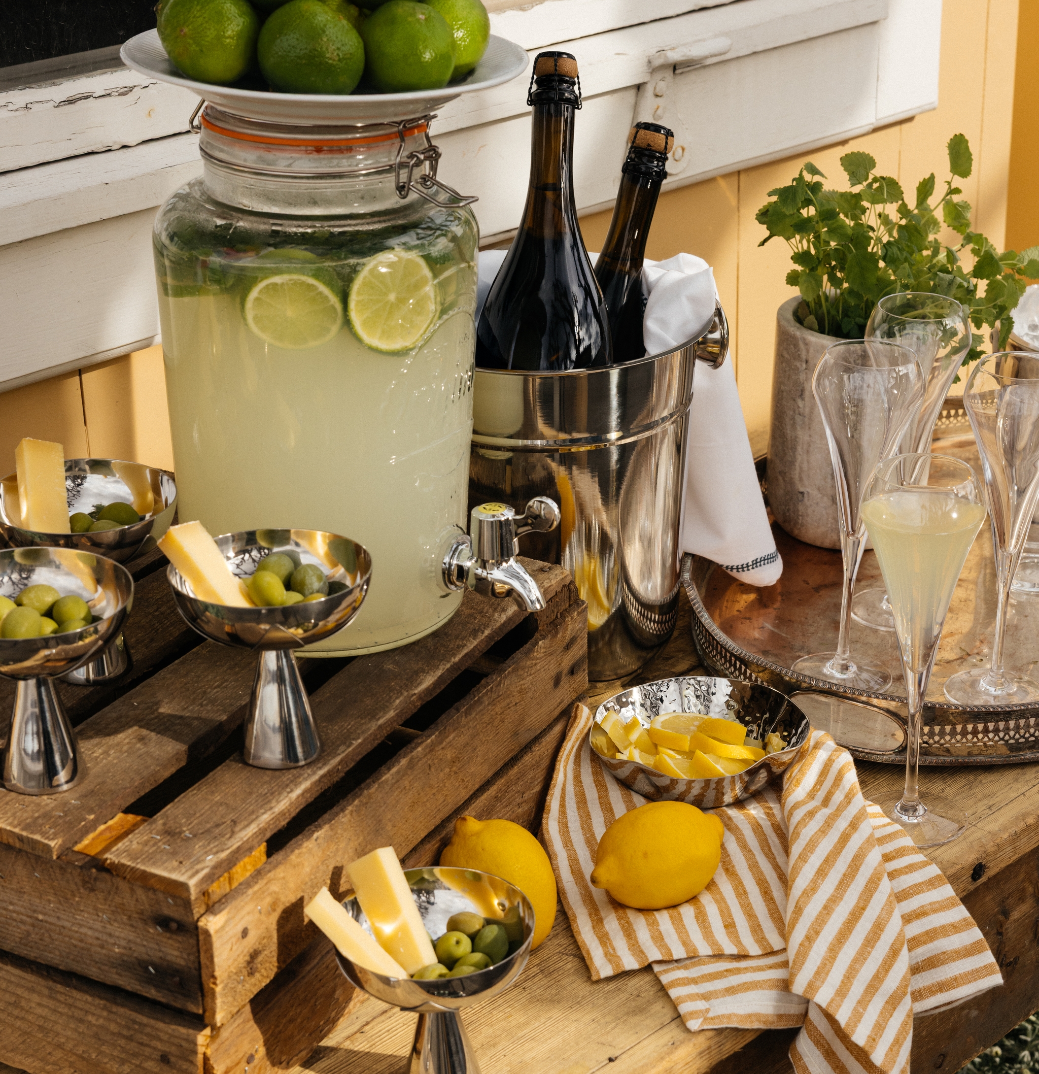 Sommerlig serveringsbord med hjemmelaget limonade, sitroner, lime, snacks og champagneglass ute på terrassen.