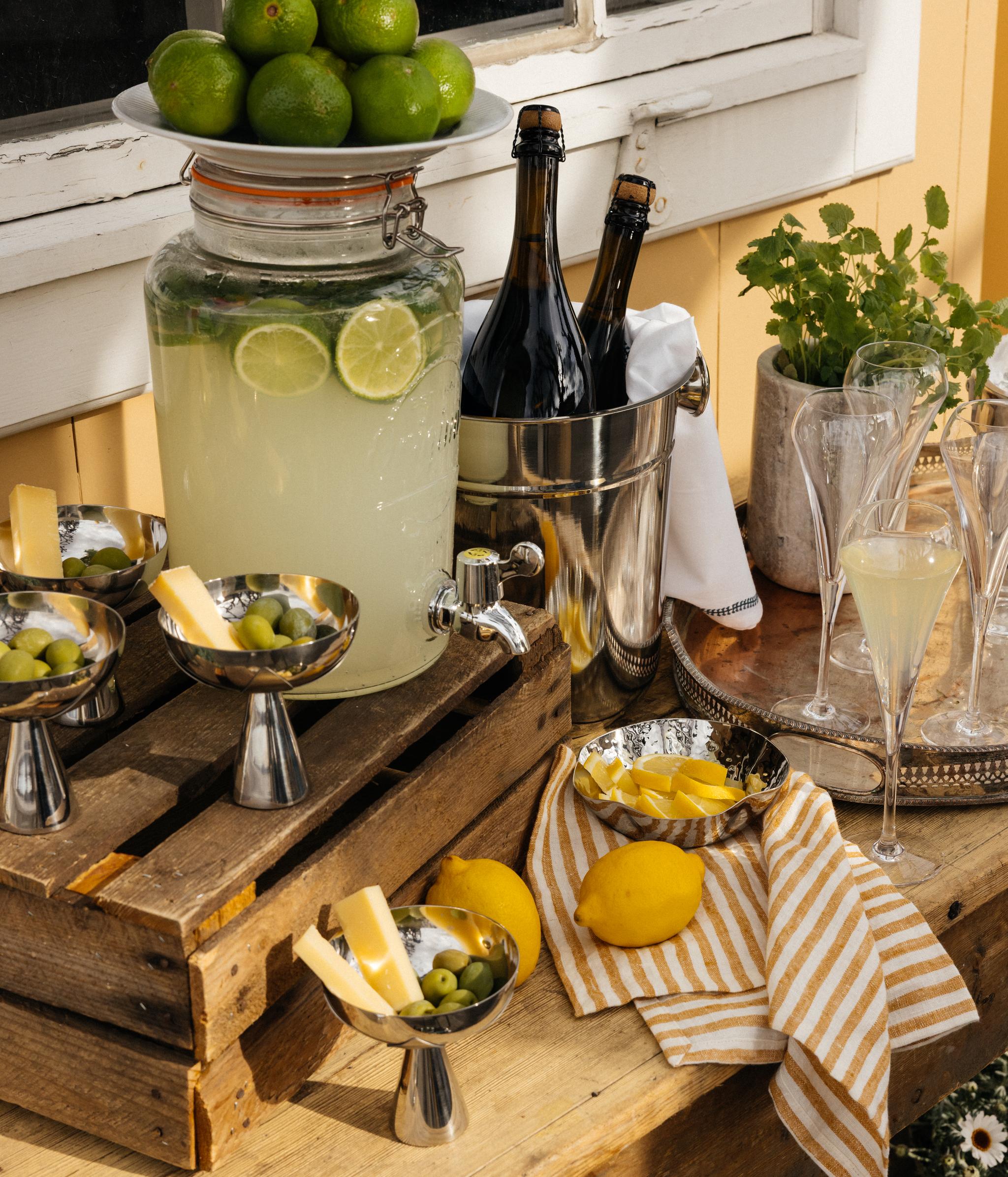 Sommerlig serveringsbord med sitronlimonade, friske sitrusfrukter, boblevin og snacks ute på terrassen.