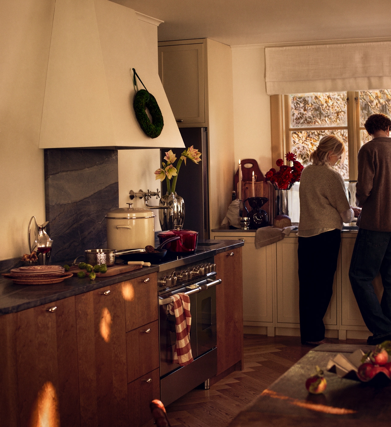 Två personer lagar mat i ett varmt, julpyntat lantkök med vedspis och vinterutsikt.