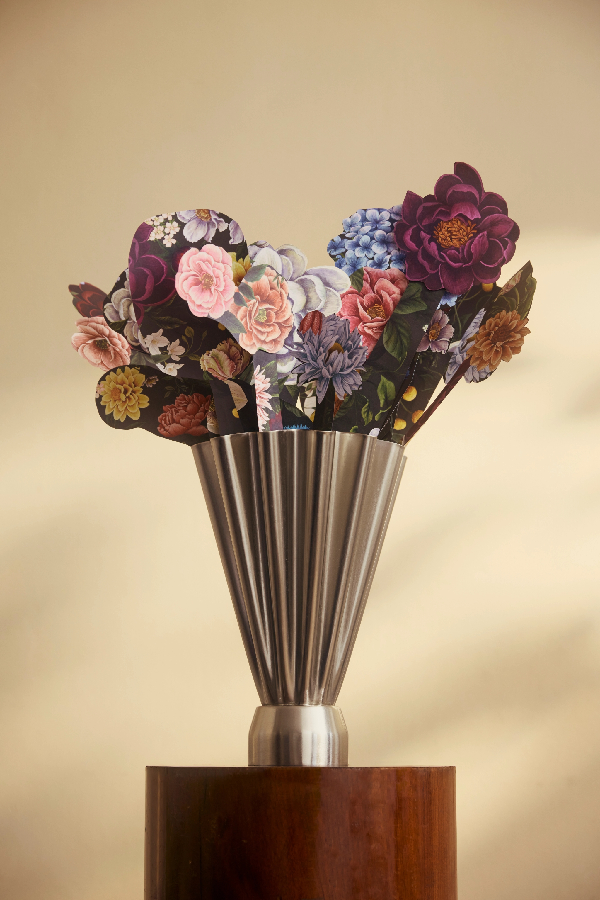 Bukett med färgglada pappersblommor i en modern metallvas på en träpiedestal mot beige bakgrund.