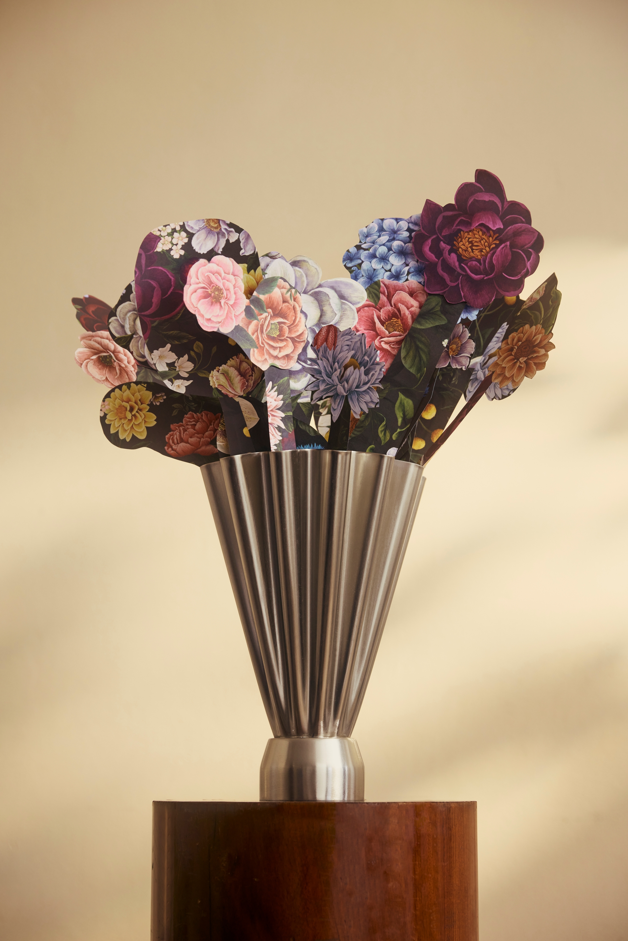 Bukett med färgglada pappersblommor i en modern metallvas på en träpiedestal mot beige bakgrund.