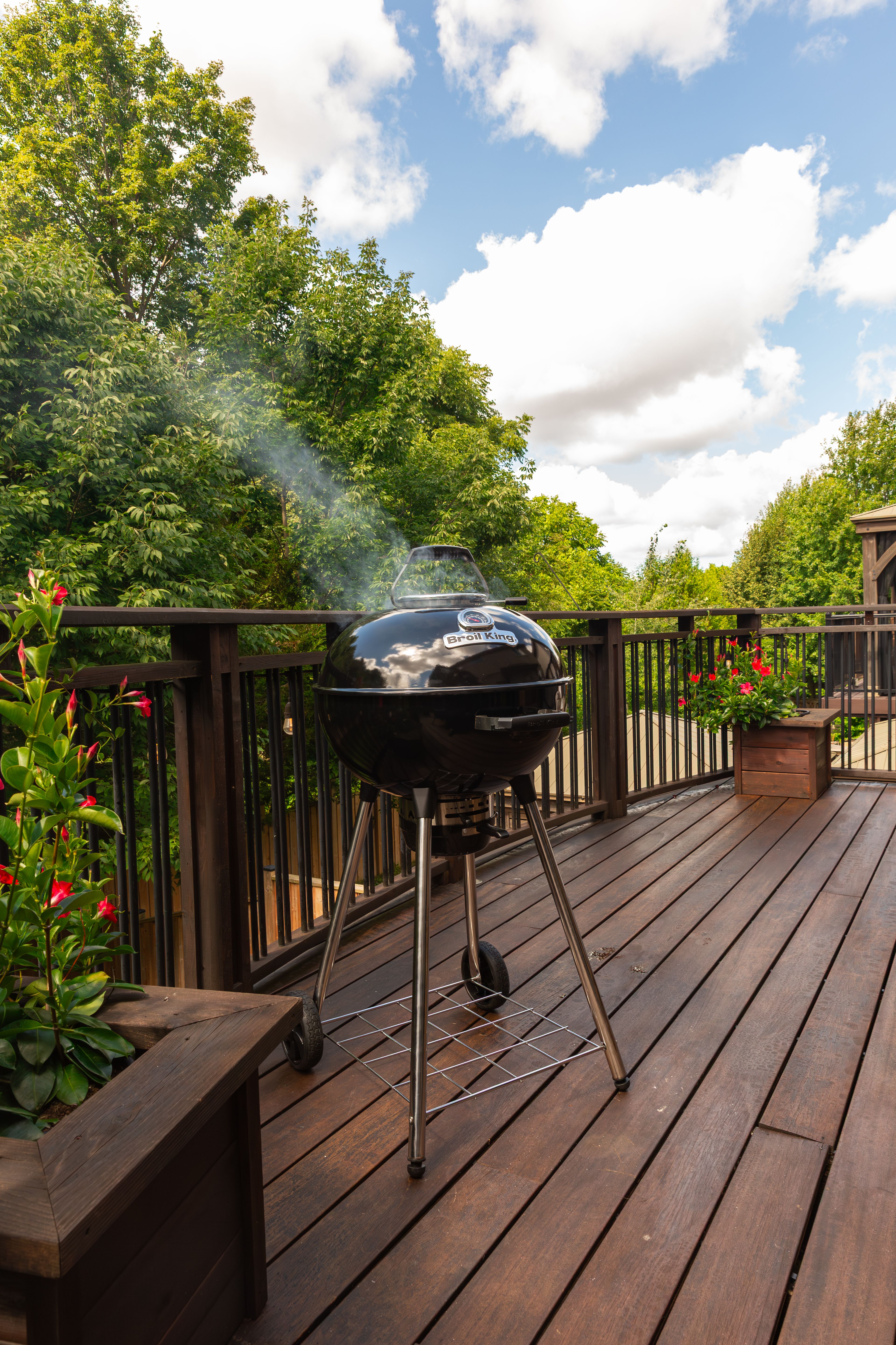 En svart klotgrill som ryker står på en träaltan, omgiven av träd och röda blommor