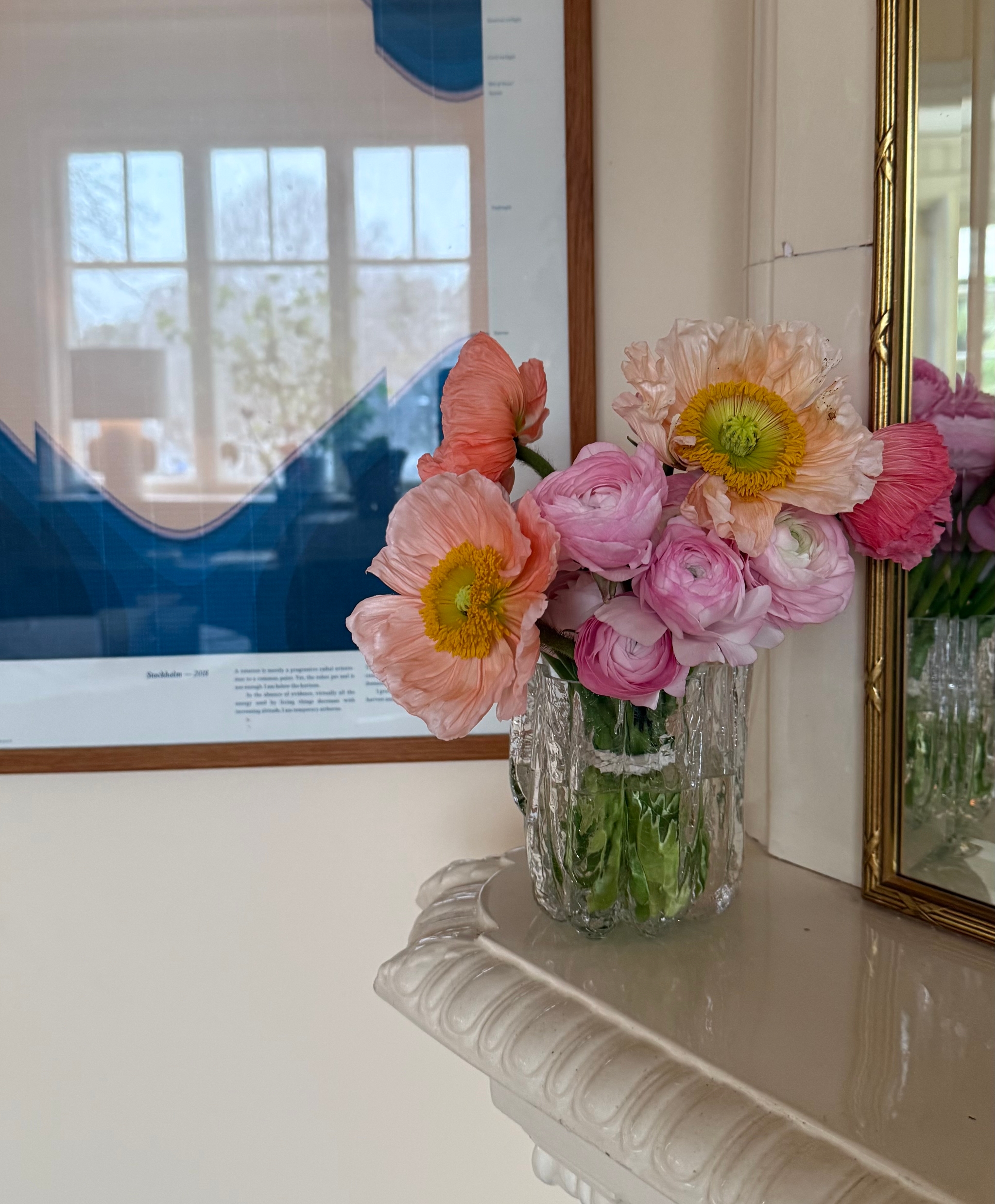 A vase of pink ranunculus and peach poppies sits on a white mantelpiece, with a blue graphic poster and a gold mirror in the background.