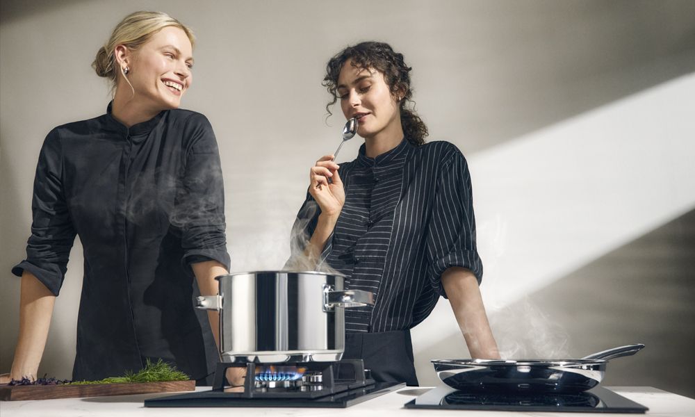 Två kockar lagar mat med en ångande gryta på en gasbrännare och en stekpanna på en induktionshäll.