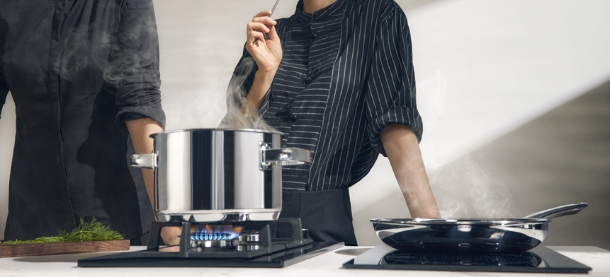 Två kockar lagar mat med en ångande gryta på en gasbrännare och en stekpanna på en induktionshäll.