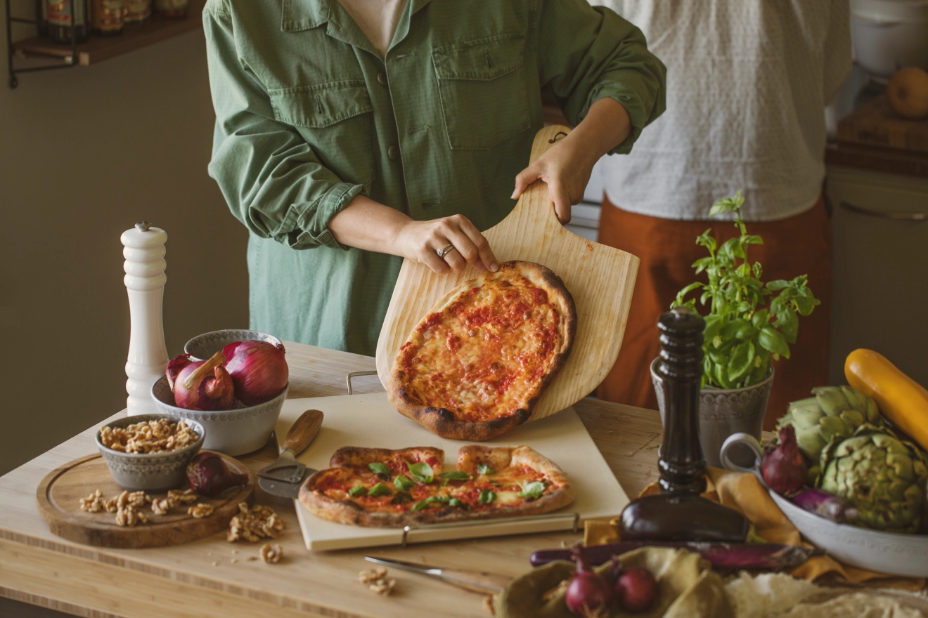 Henkilö nostaa vastapaistetun kotitekoisen pizzan puulastalla keittiön pöydälle, joka on täynnä tuoreita vihanneksia ja yrttejä.