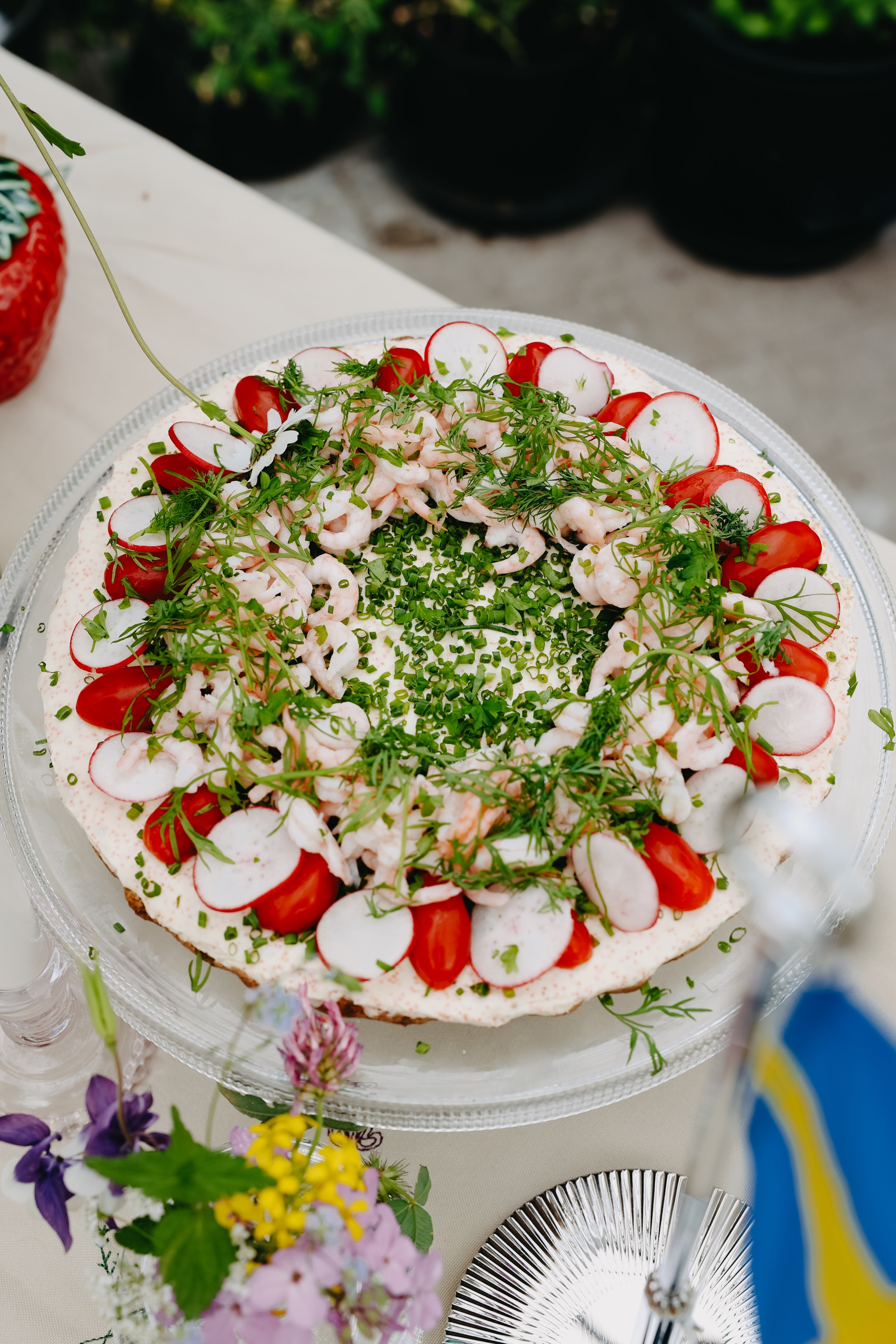 En festlig svensk smörgåstårta toppad med räkor, rädisor, körsbärstomater och färska örter på ett dukat bord.