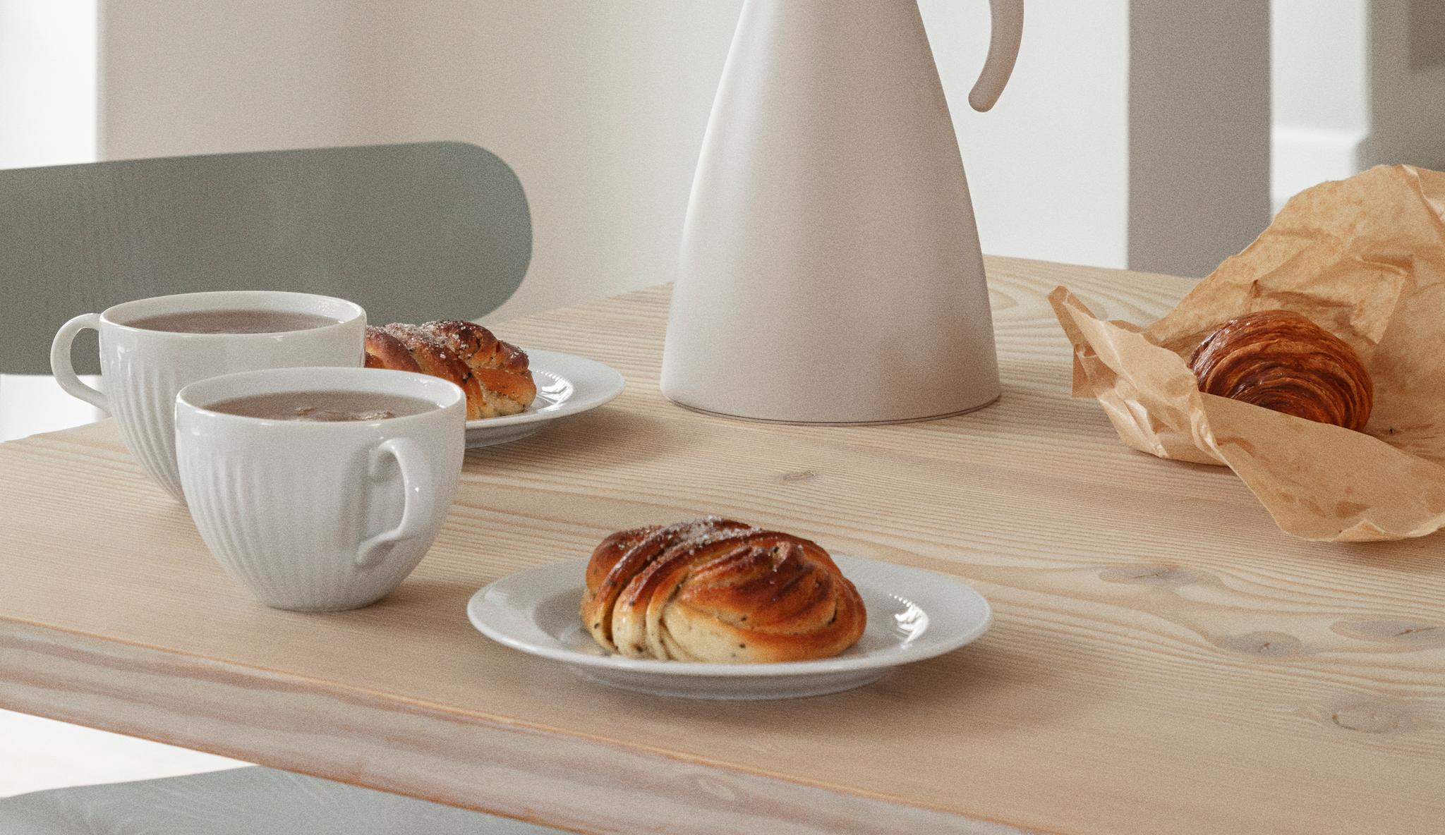 Vaalealle puupöydälle katettu kahvihetki, jossa on valkoinen kannu, kahvikuppeja, korvapuusteja ja croissant.