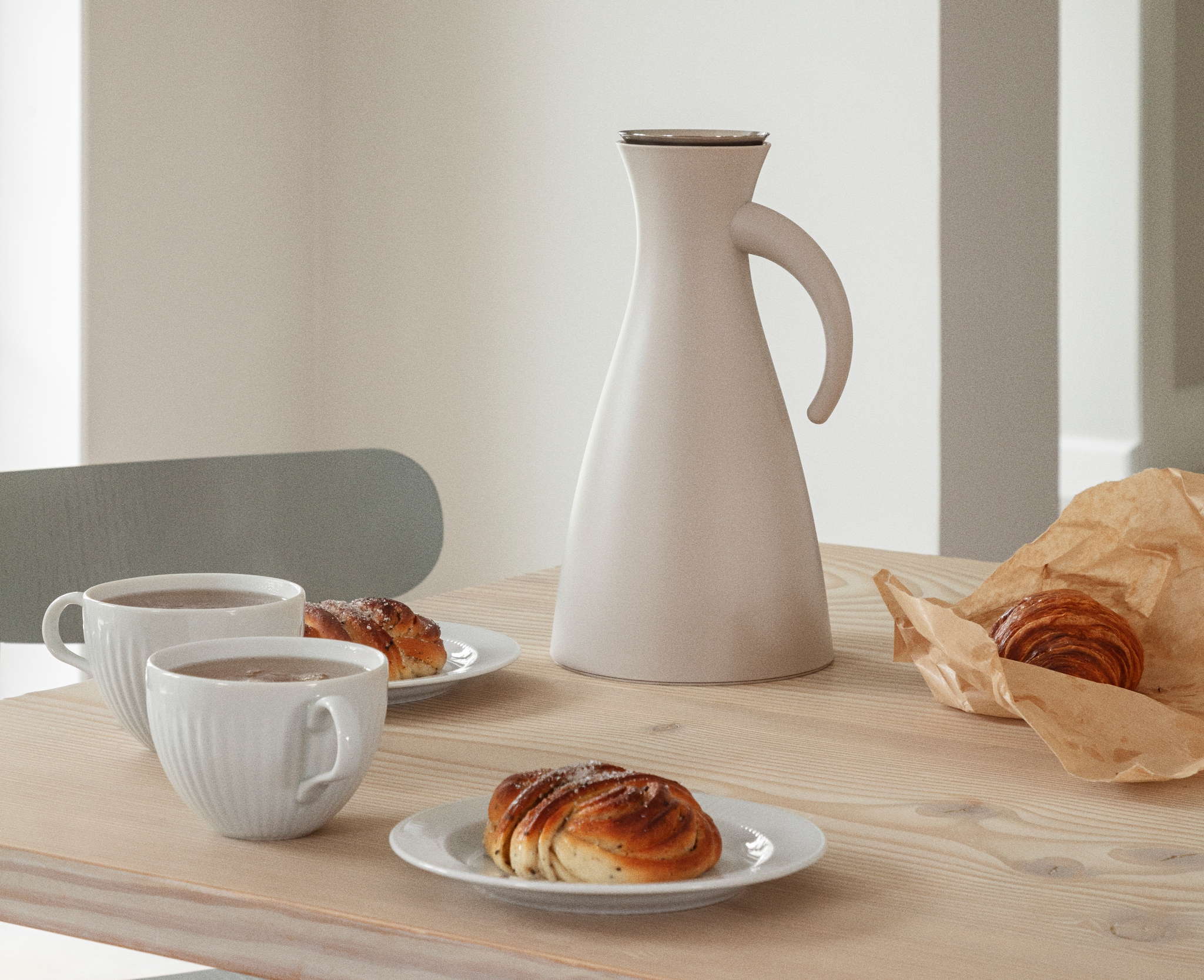Vaalealla puupöydällä on kahvikuppeja, valkoinen kannu sekä lautasilla tuoreita korvapuusteja ja croissant.