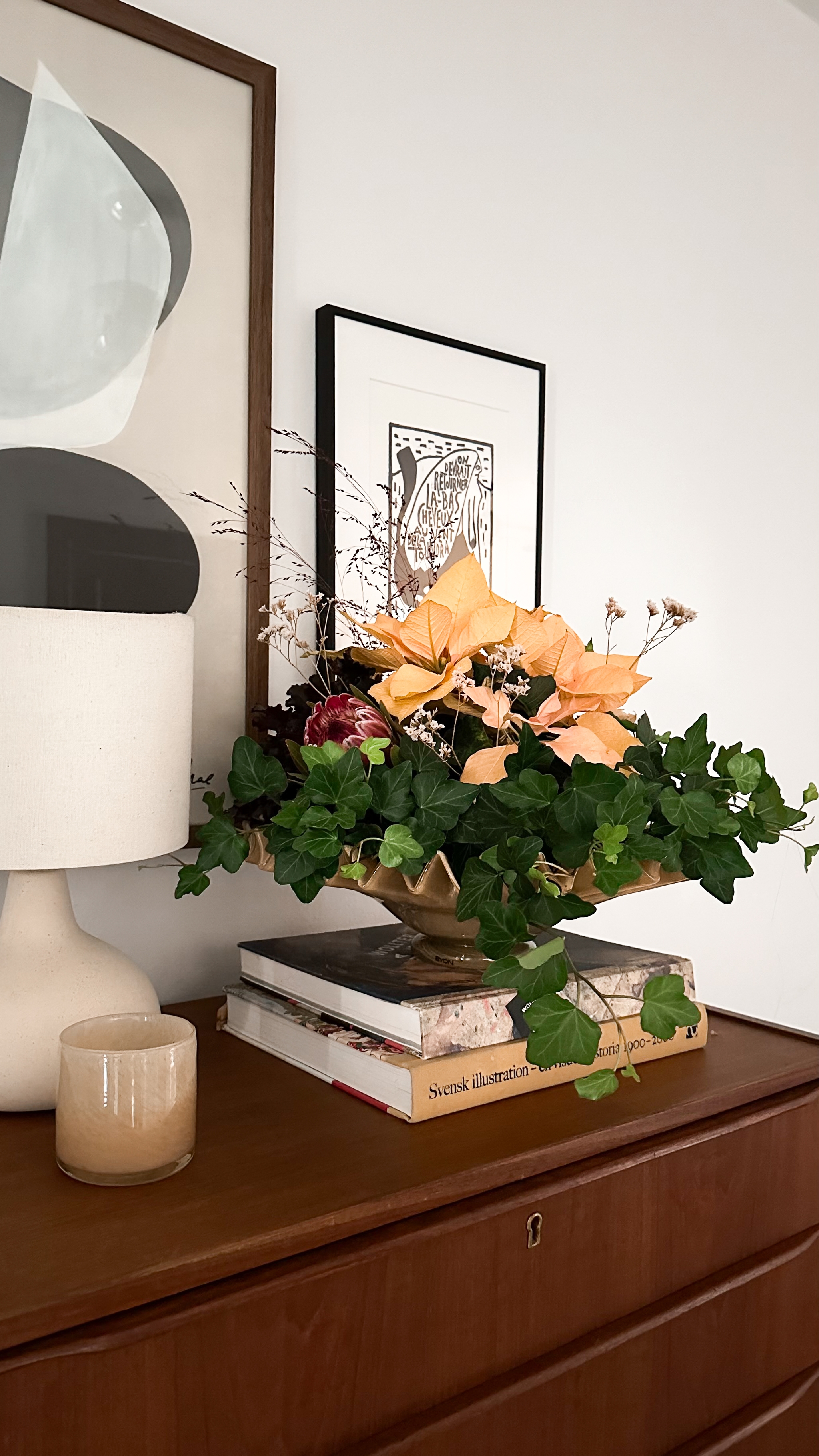 A floral arrangement with peach flowers and green ivy in a gold wavy bowl sits on books atop a wooden dresser, with framed art and a lamp in the background.