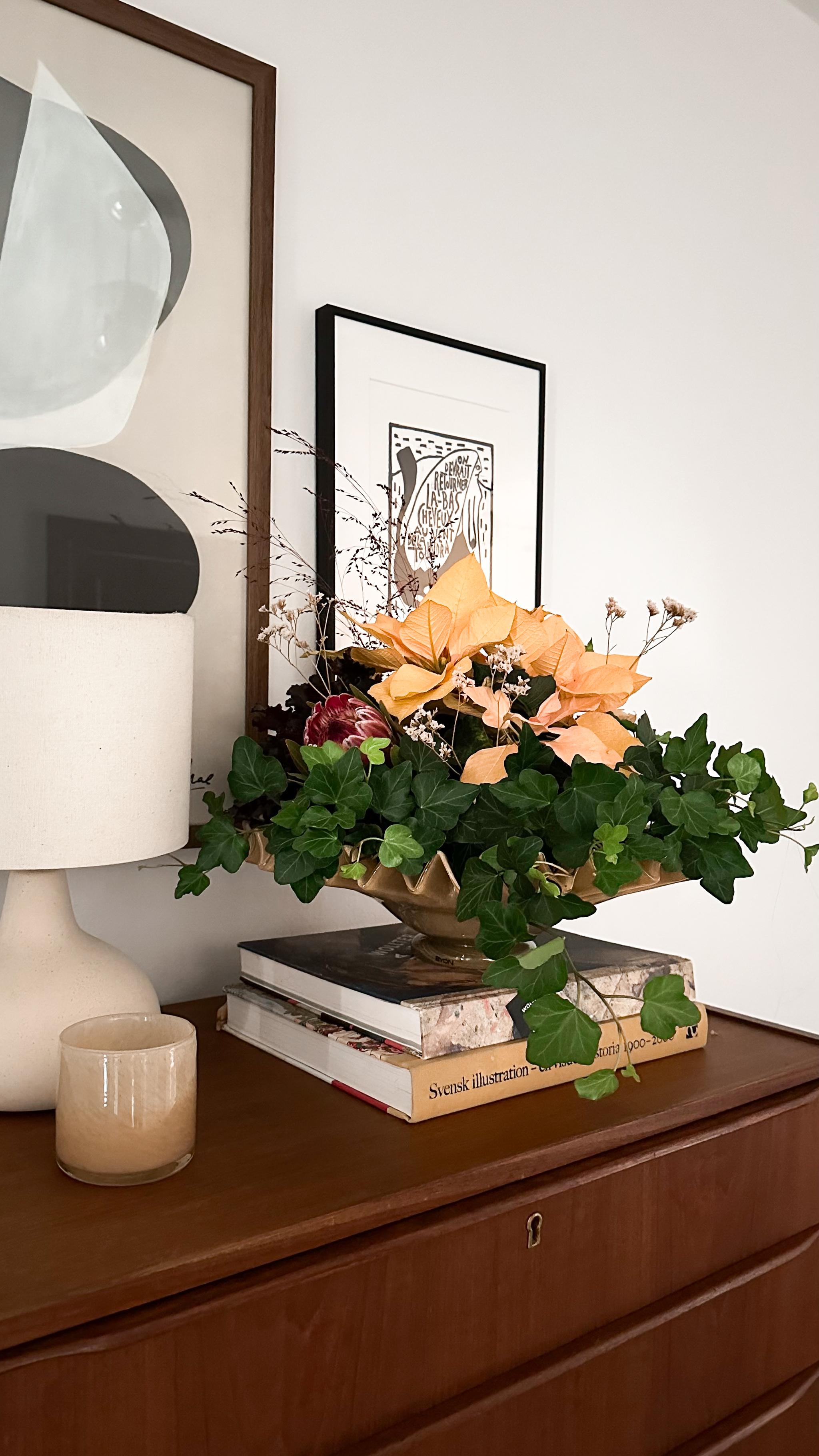 A floral arrangement with peach flowers and green ivy in a gold wavy bowl sits on books atop a wooden dresser, with framed art and a lamp in the background.