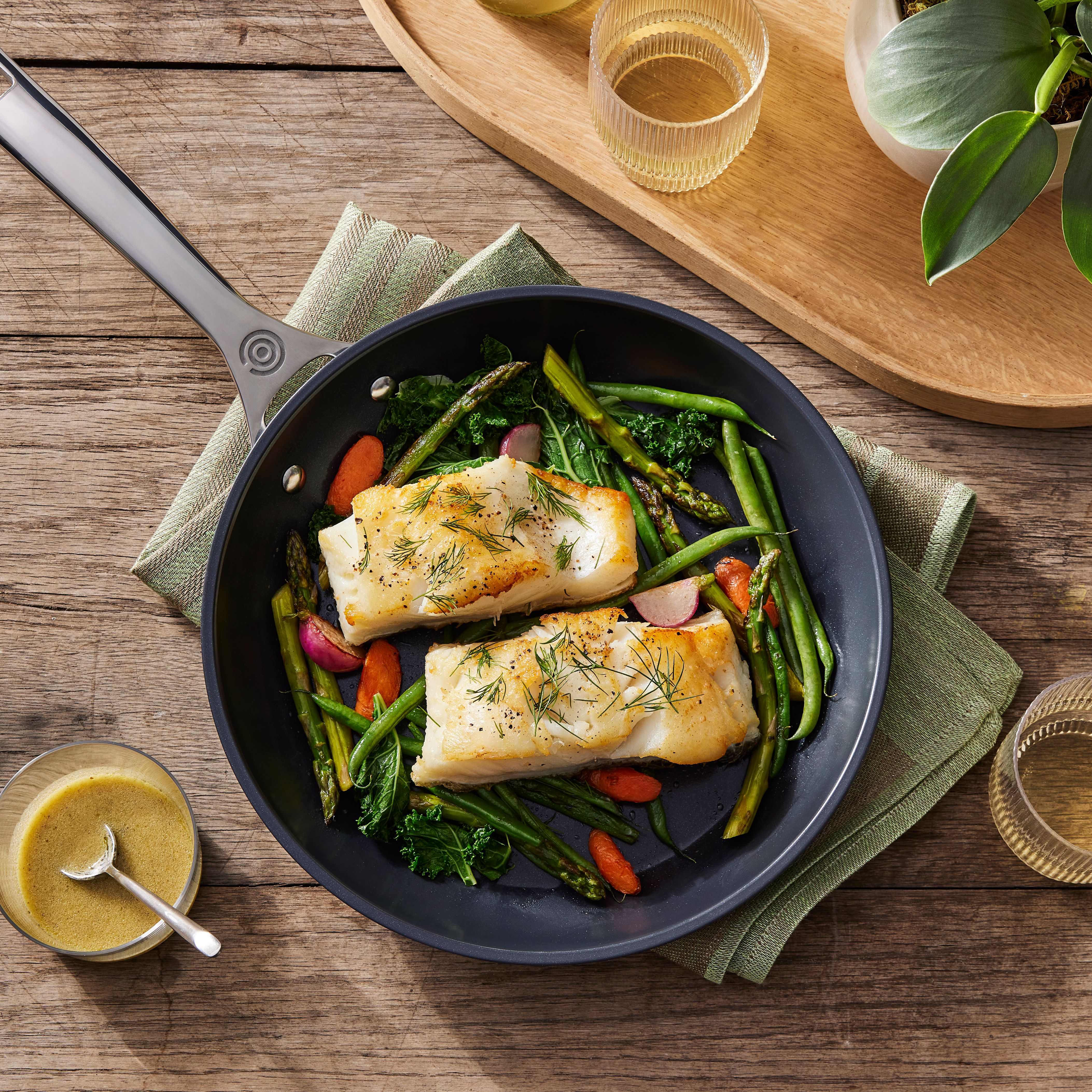 En mörk stekpanna innehåller två stekta vita fiskfiléer toppade med dill och peppar, omgivna av sparris, gröna bönor, grönkål, morötter och rädisor, allt på ett träbord.
